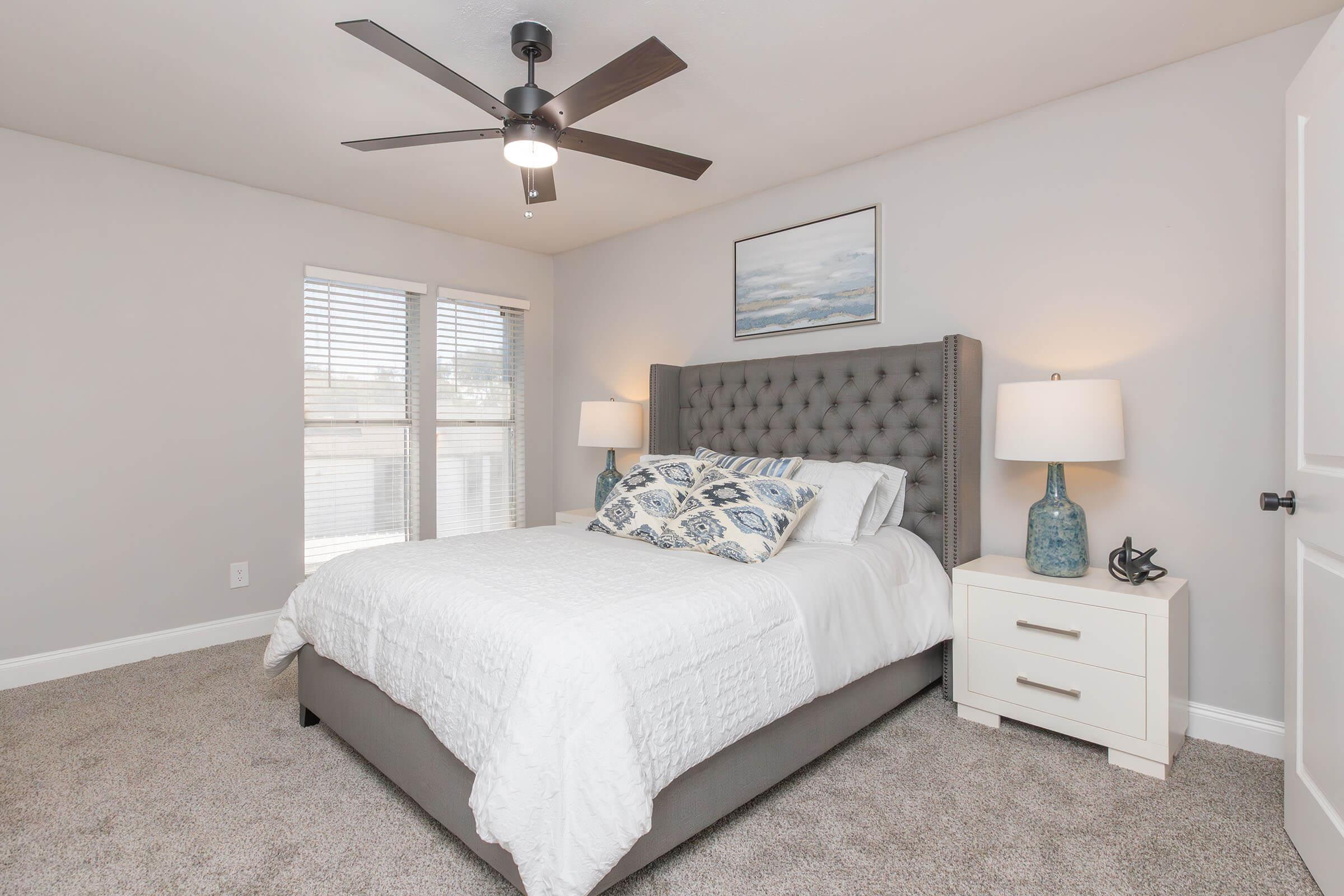 A cozy bedroom featuring a king-size bed with a tufted gray headboard, decorative pillows, and crisp white bedding. Two bedside lamps illuminate a minimalist nightstand. Soft gray walls and large windows let in natural light, complemented by a ceiling fan and a serene landscape painting on the wall.