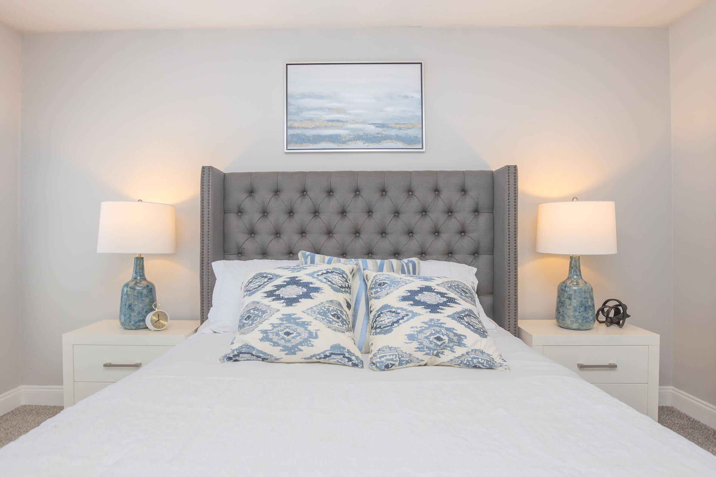 A cozy bedroom featuring a gray tufted headboard, white bedding with decorative blue and white pillows, and two bedside tables with blue lamps. A piece of abstract art hangs above the bed, and soft gray walls create a calming atmosphere.