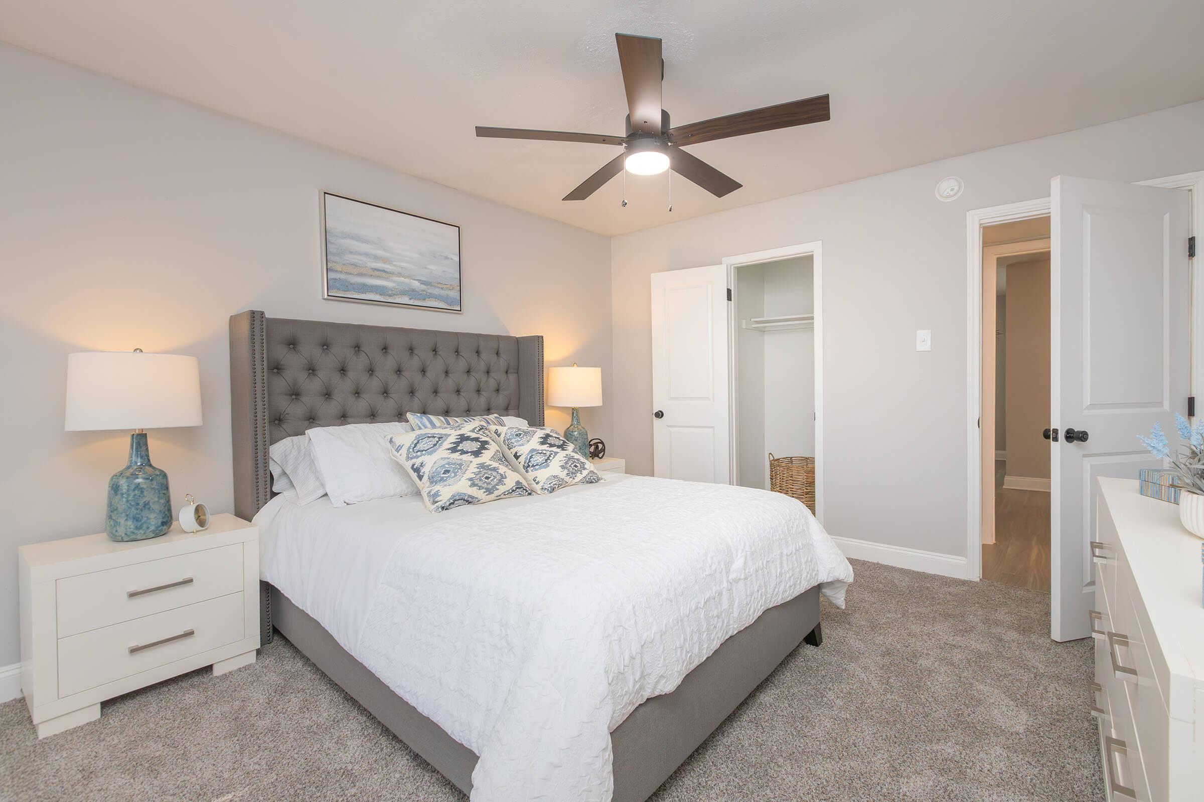 A cozy bedroom featuring a gray upholstered bed with white bedding and decorative pillows. There are bedside lamps on white nightstands, a ceiling fan above, and a close closet with an open door. The walls are painted light gray, and there's a painting hanging above the bed.