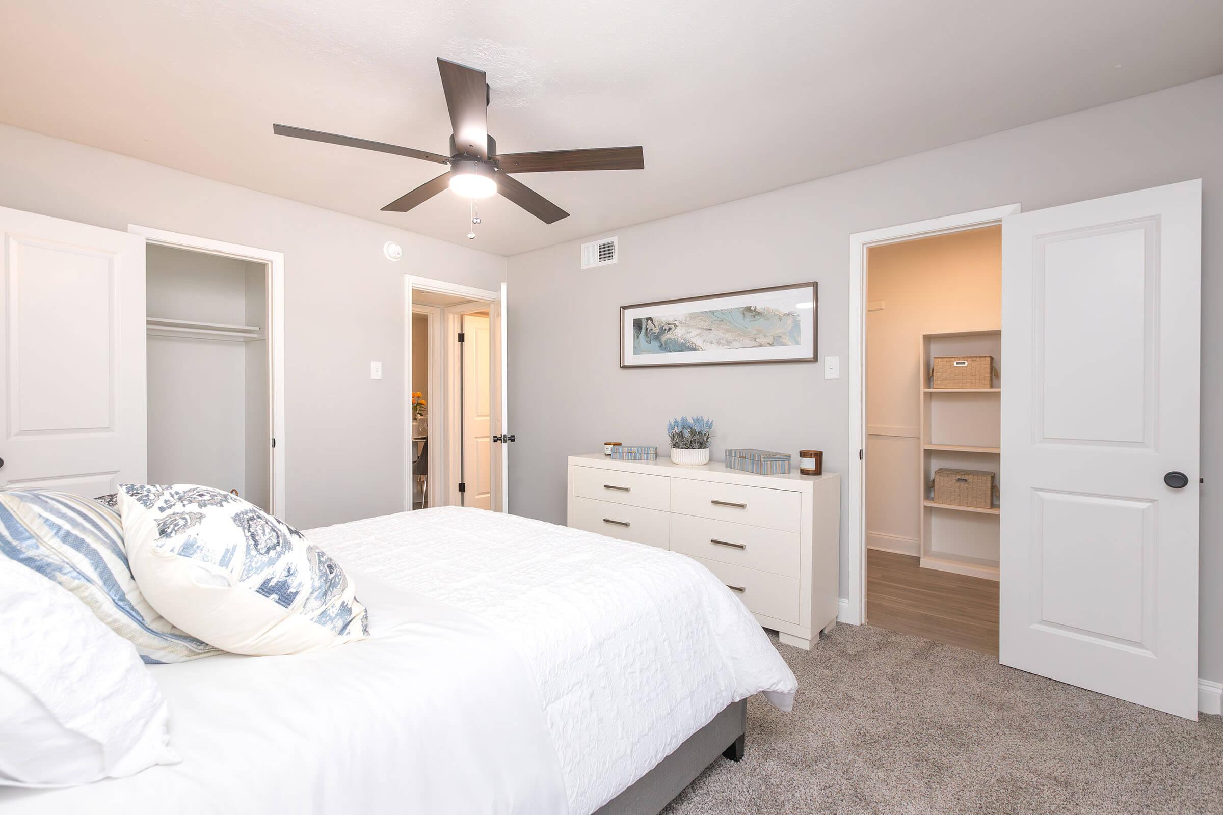 A modern bedroom featuring a white bed with decorative pillows, a light-gray wall, and a ceiling fan. There's a white dresser against the wall, and two doors leading to closets and additional space. Soft carpet underfoot completes the cozy atmosphere.