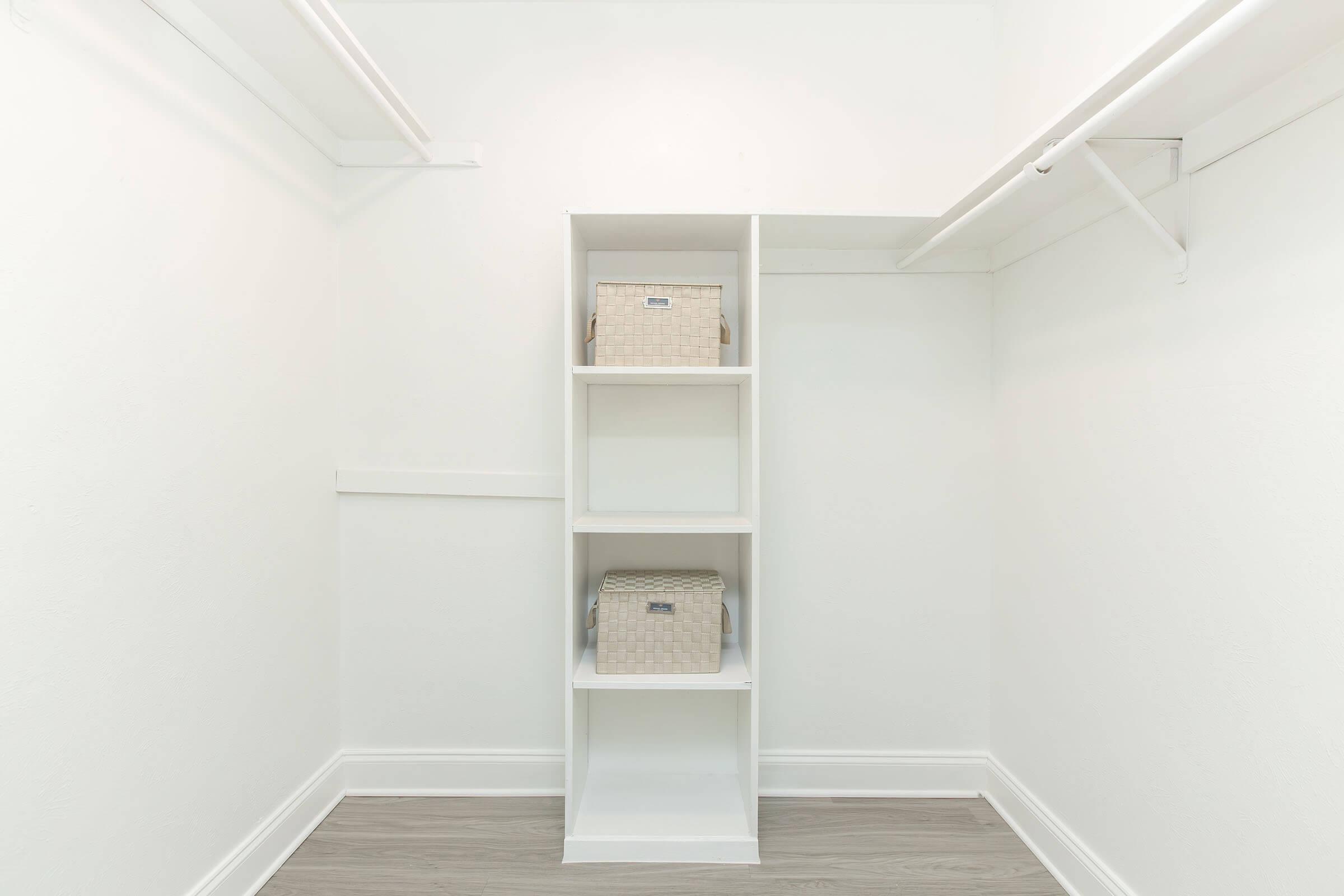 A neatly organized closet featuring white walls and a light wooden floor. The closet has a tall white shelving unit with three sections, two of which contain storage boxes. The space appears bright and minimalist, conveying a sense of cleanliness and order.