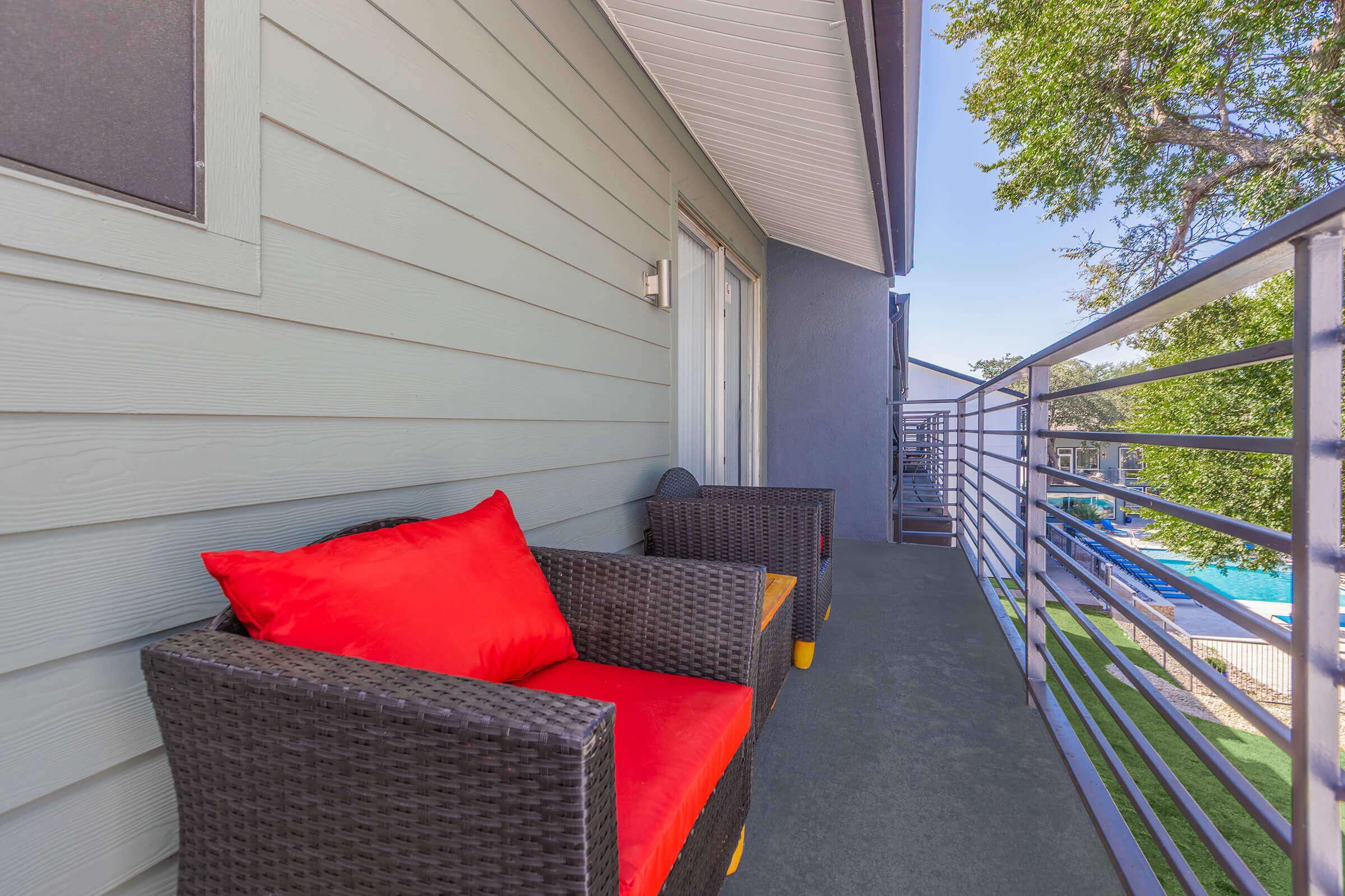 A balcony featuring two wicker chairs with bright red cushions. The flooring is dark and smooth, and there is a railing that overlooks a pool area and greenery, creating a relaxing outdoor space.