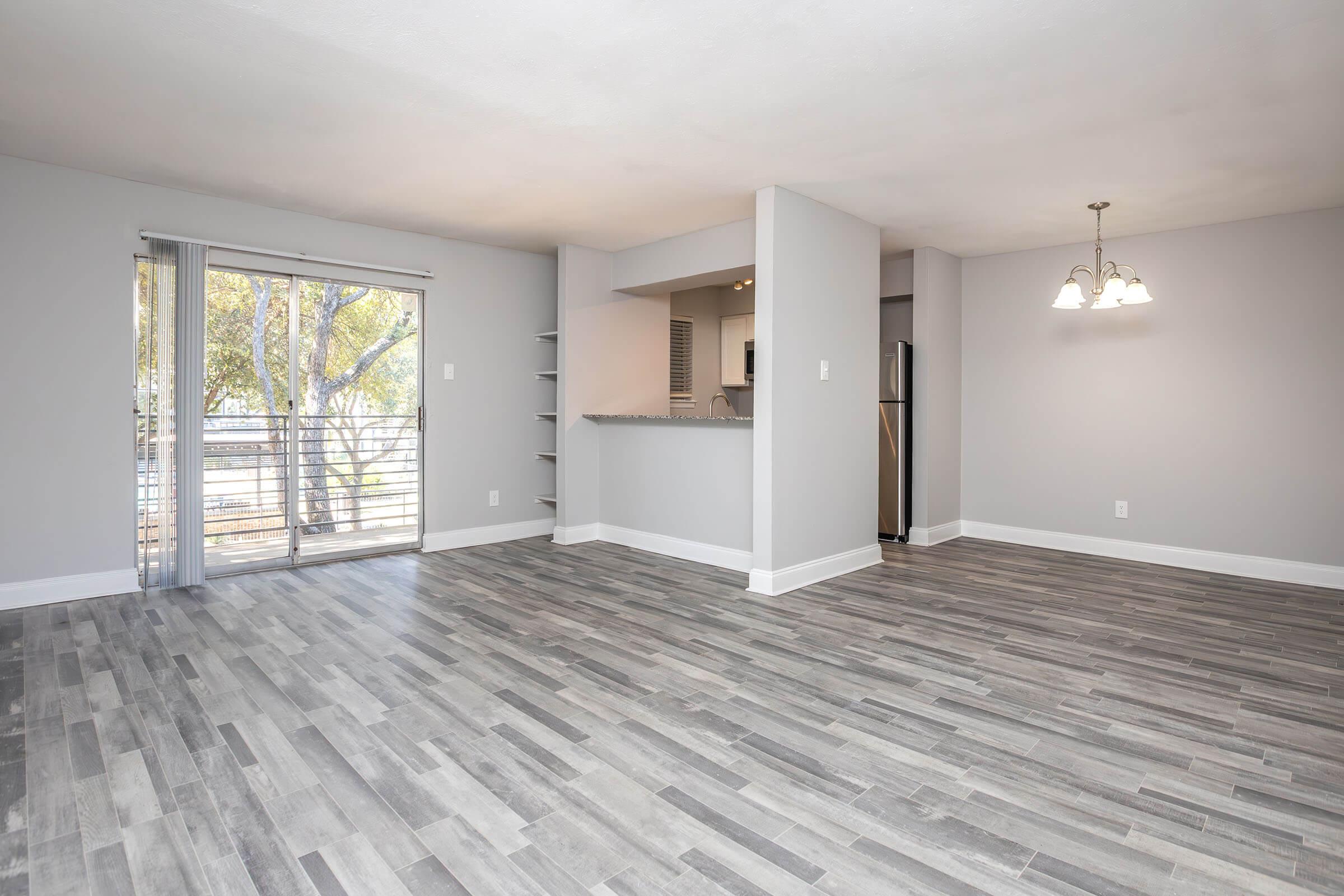 A spacious, light-filled living area featuring gray walls, modern laminate flooring, and a sliding glass door leading to a balcony. The open floor plan includes a countertop bar area and adjacent kitchen with stainless steel appliances visible in the background. Bright lighting from a hanging fixture adds warmth.
