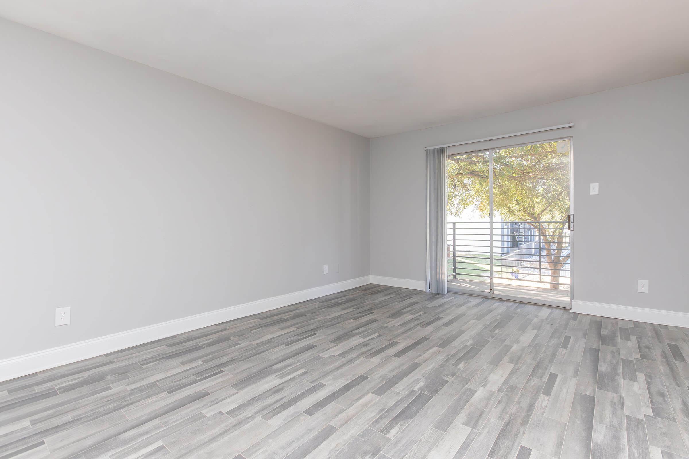 A spacious, empty room with gray walls and a light gray wood-like floor. There is a large sliding glass door leading to an outdoor area with trees visible outside. The room has minimal decor, providing a clean and modern aesthetic.