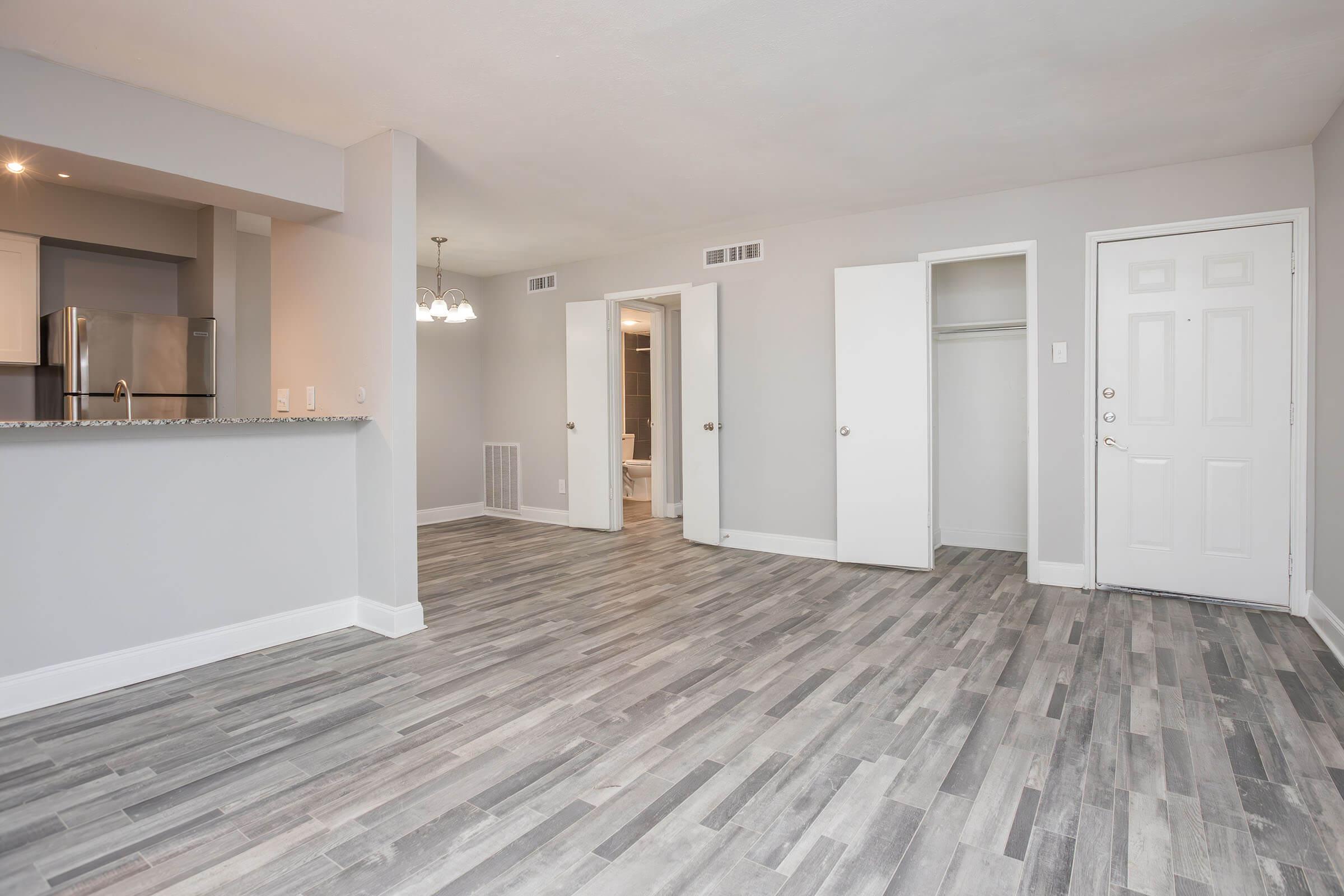 Spacious interior of an apartment featuring gray laminate flooring, a modern kitchen with stainless steel appliances visible in the background, and a well-lit area with doorways leading to closets and a bathroom. The walls are painted in a light neutral color, creating a bright and open atmosphere.