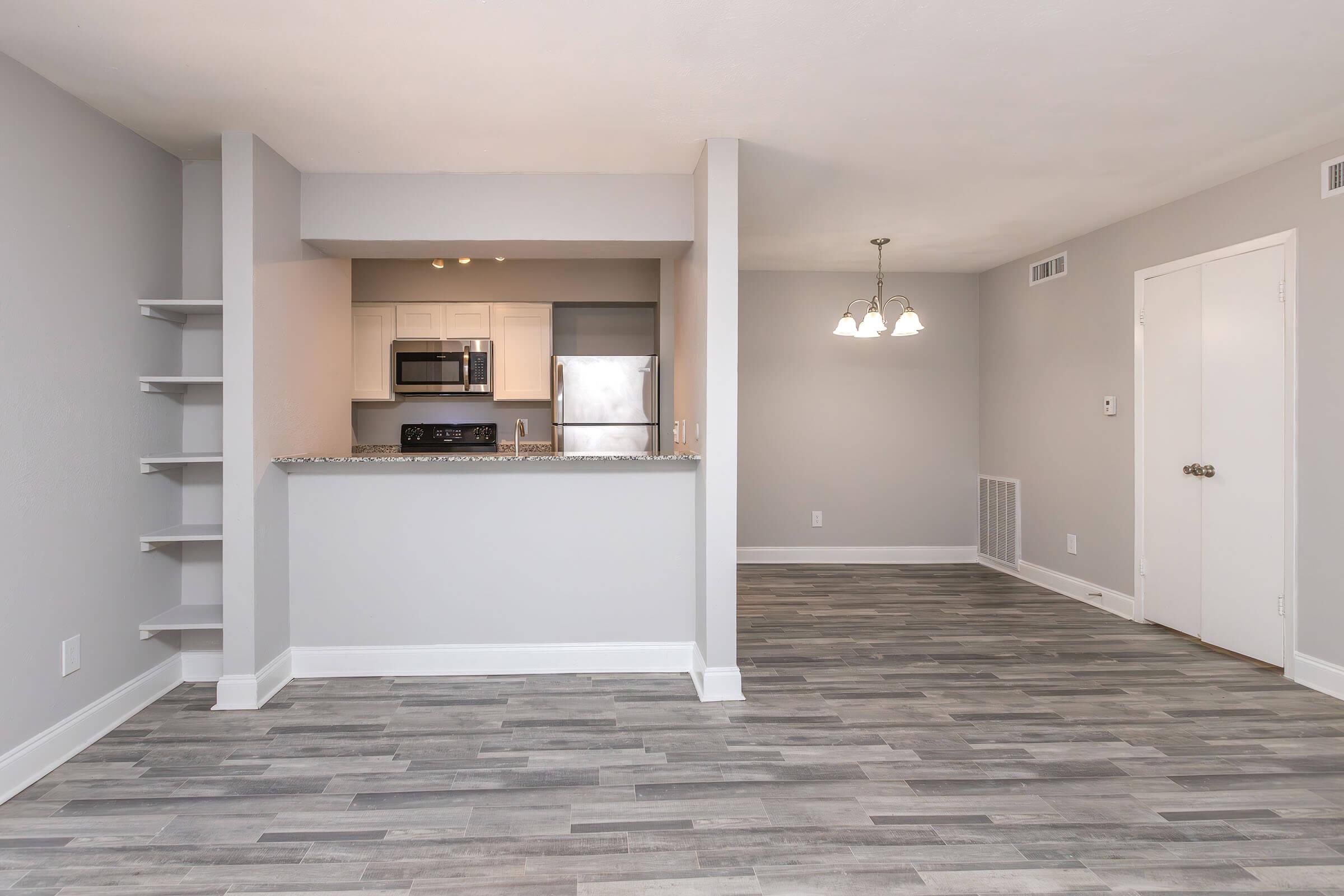 Modern open-concept living space featuring a kitchen with stainless steel appliances, a granite countertop, and light gray cabinets. The dining area includes a chandelier, and the flooring is a stylish gray laminate. Neutral wall colors enhance the spacious and inviting atmosphere.