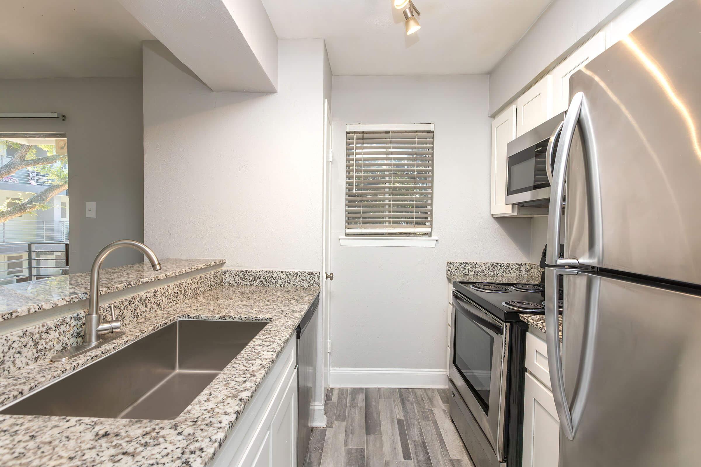 A modern kitchen featuring stainless steel appliances, including a refrigerator, oven, and microwave. The countertops are made of granite, and there's a double sink. Natural light enters through a small window with blinds, illuminating the space. The flooring is a light wood look.