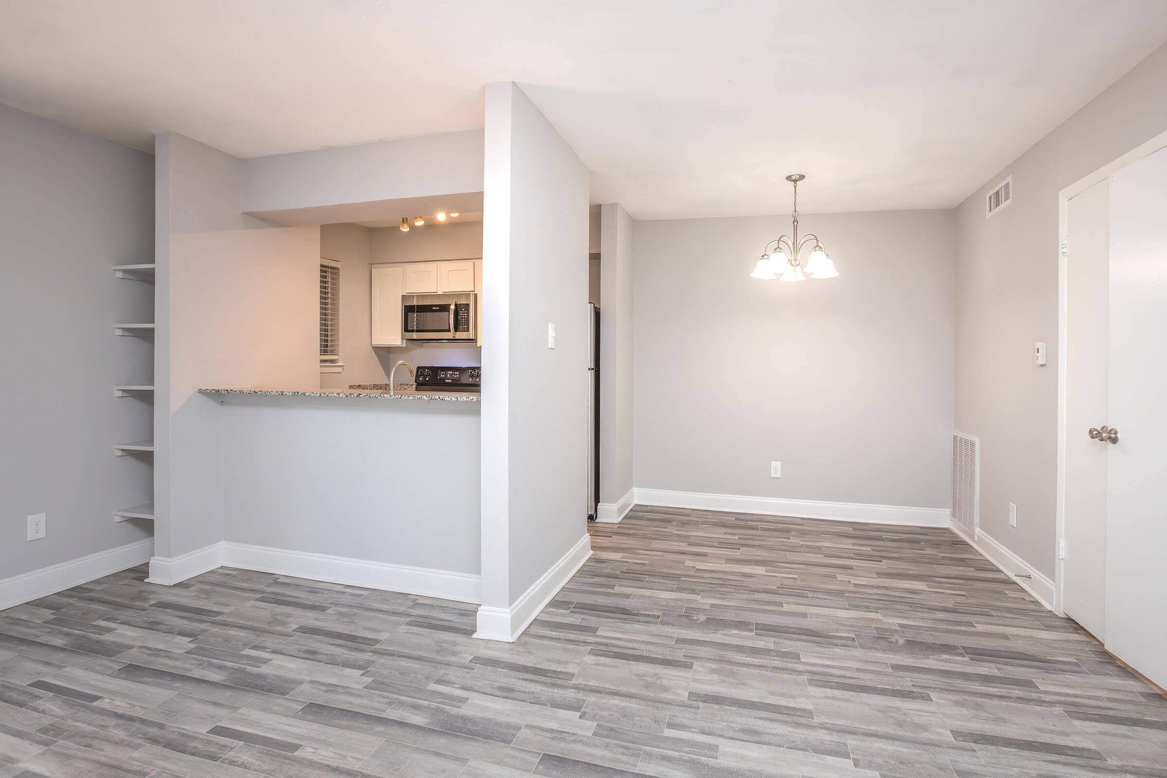 A modern, open-concept living space featuring a kitchen with granite countertops, white cabinetry, and stainless steel appliances. The dining area is illuminated by a chandelier, and the flooring is a light wood-look tile. The walls are painted in a soft gray, adding to the contemporary feel of the room.