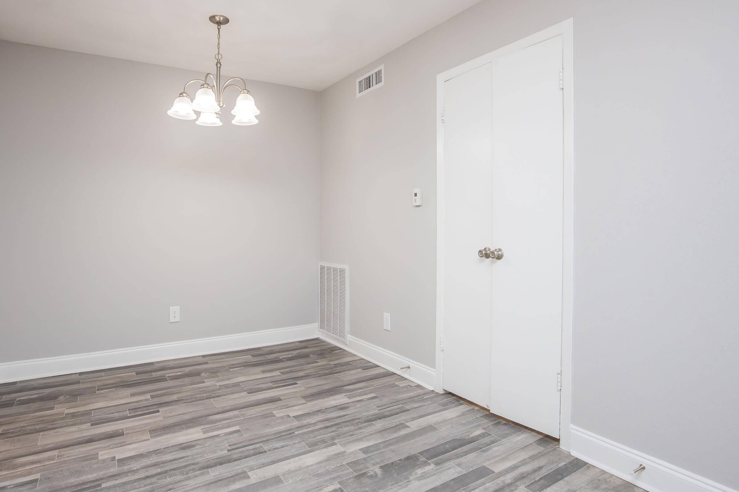 An empty room with gray walls and a light fixture featuring three bulbs. The floor has a wood-like pattern in shades of gray. There is a closed white door on the back wall and a small air vent near the floor. The room is well-lit and appears to be newly renovated.