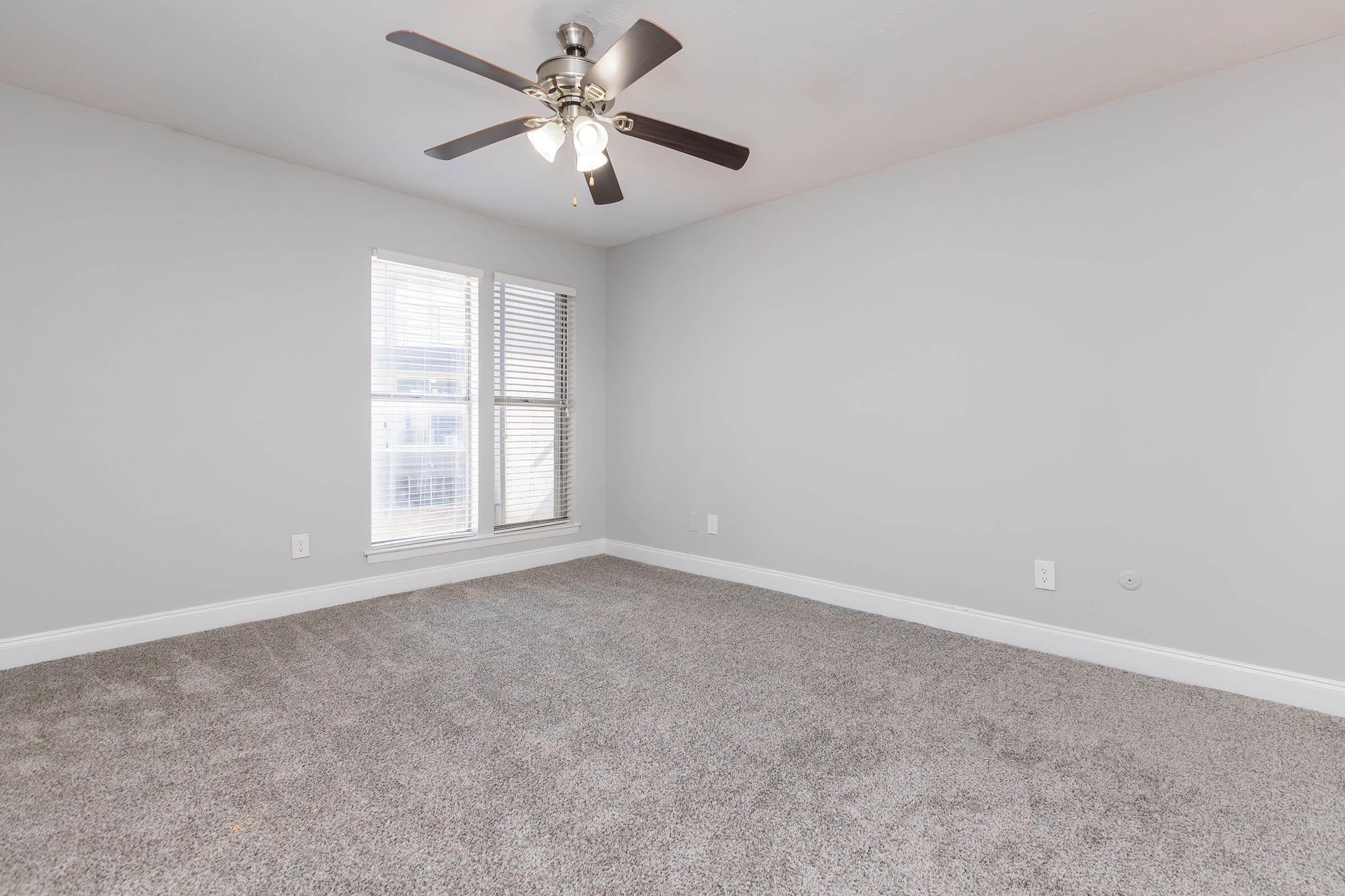 A bright, empty room featuring light gray walls and a plush carpet. A ceiling fan with three lights hangs from the ceiling, and two windows with white blinds allow natural light to enter the space. The room is ready for decorating or furnishing.