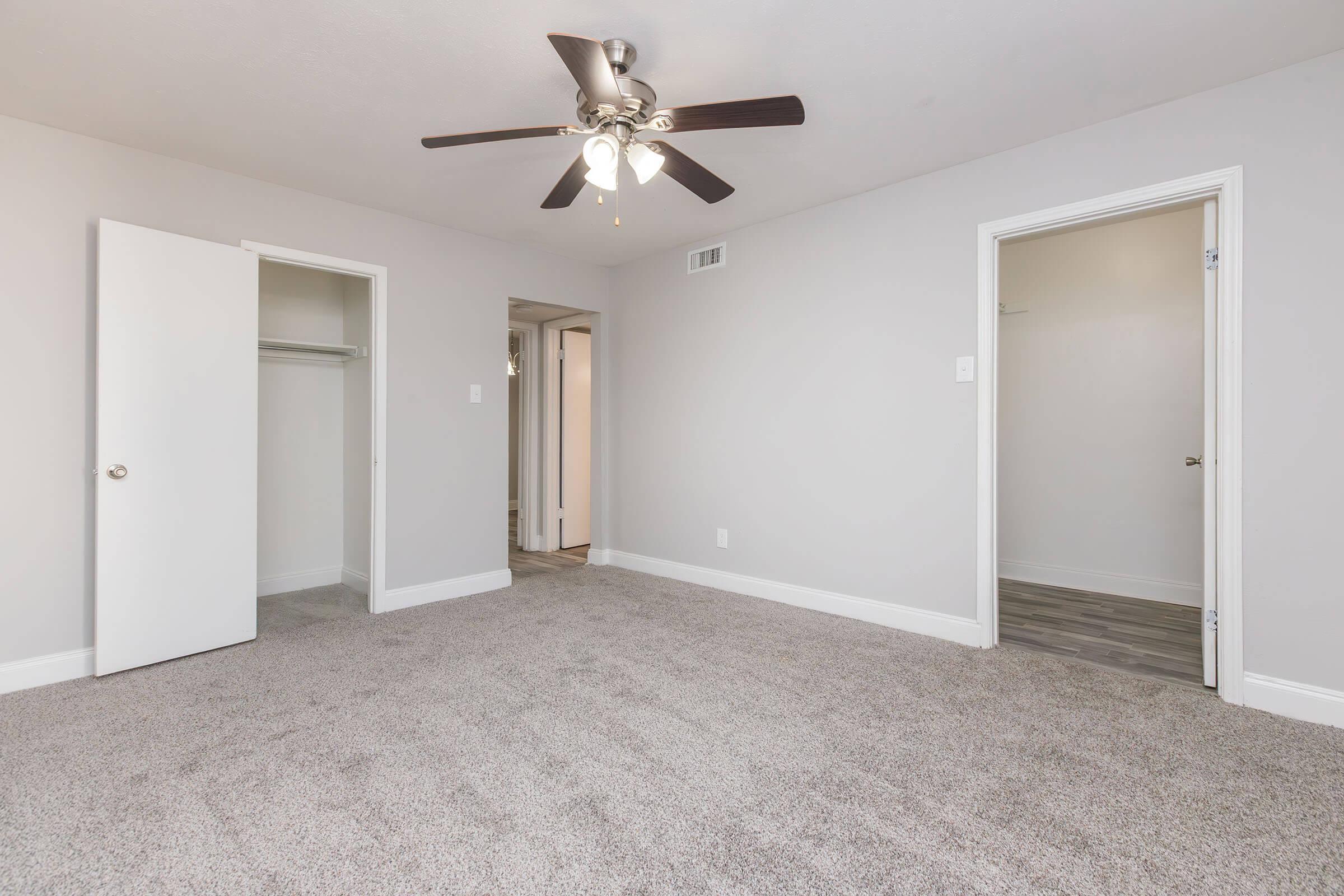 A well-lit, empty bedroom featuring light gray walls, plush carpet flooring, and a ceiling fan. There are two closed doors, one leading to a closet and the other to what appears to be a hallway. The room’s neutral color palette creates a spacious feel.