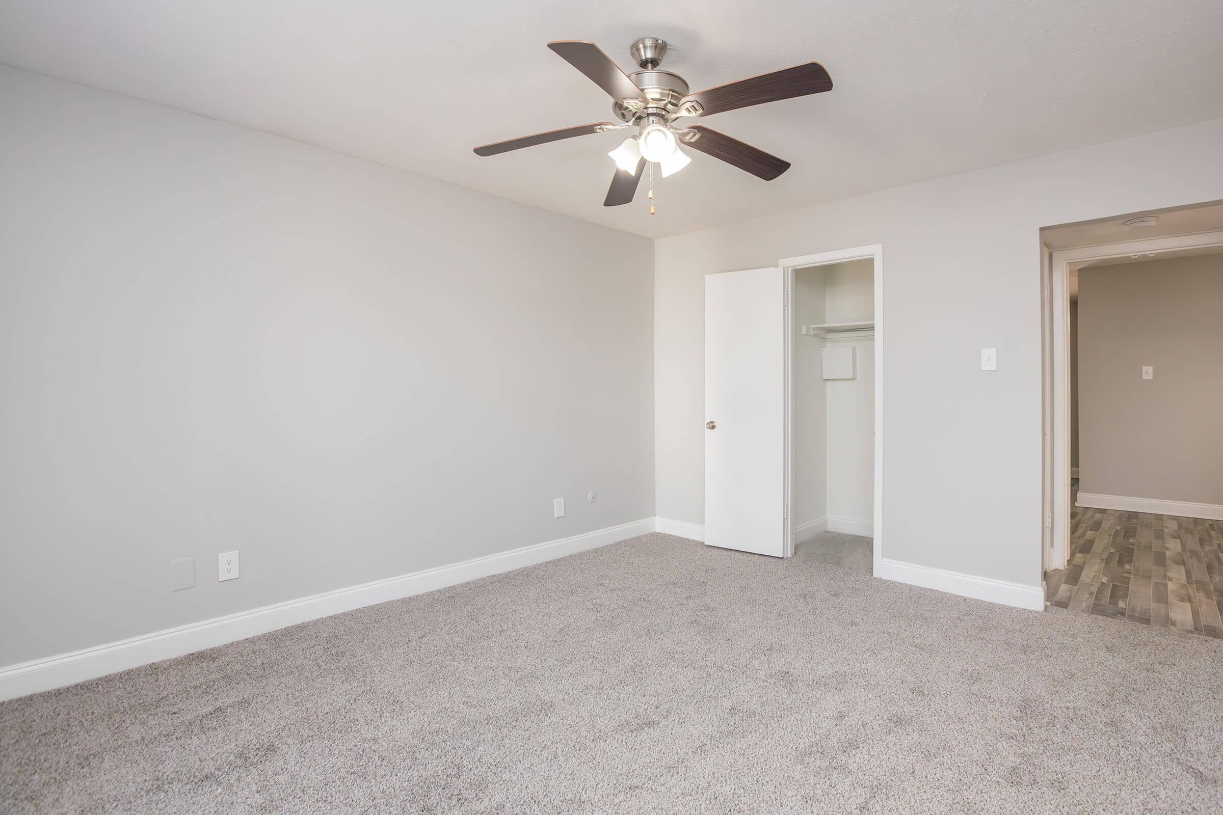 A spacious, empty bedroom featuring soft beige carpet, light gray walls, and a ceiling fan with three lights. There's a white closet with a sliding door on the left and an open doorway leading to another area on the right. The room is well-lit, offering a clean and neutral aesthetic.