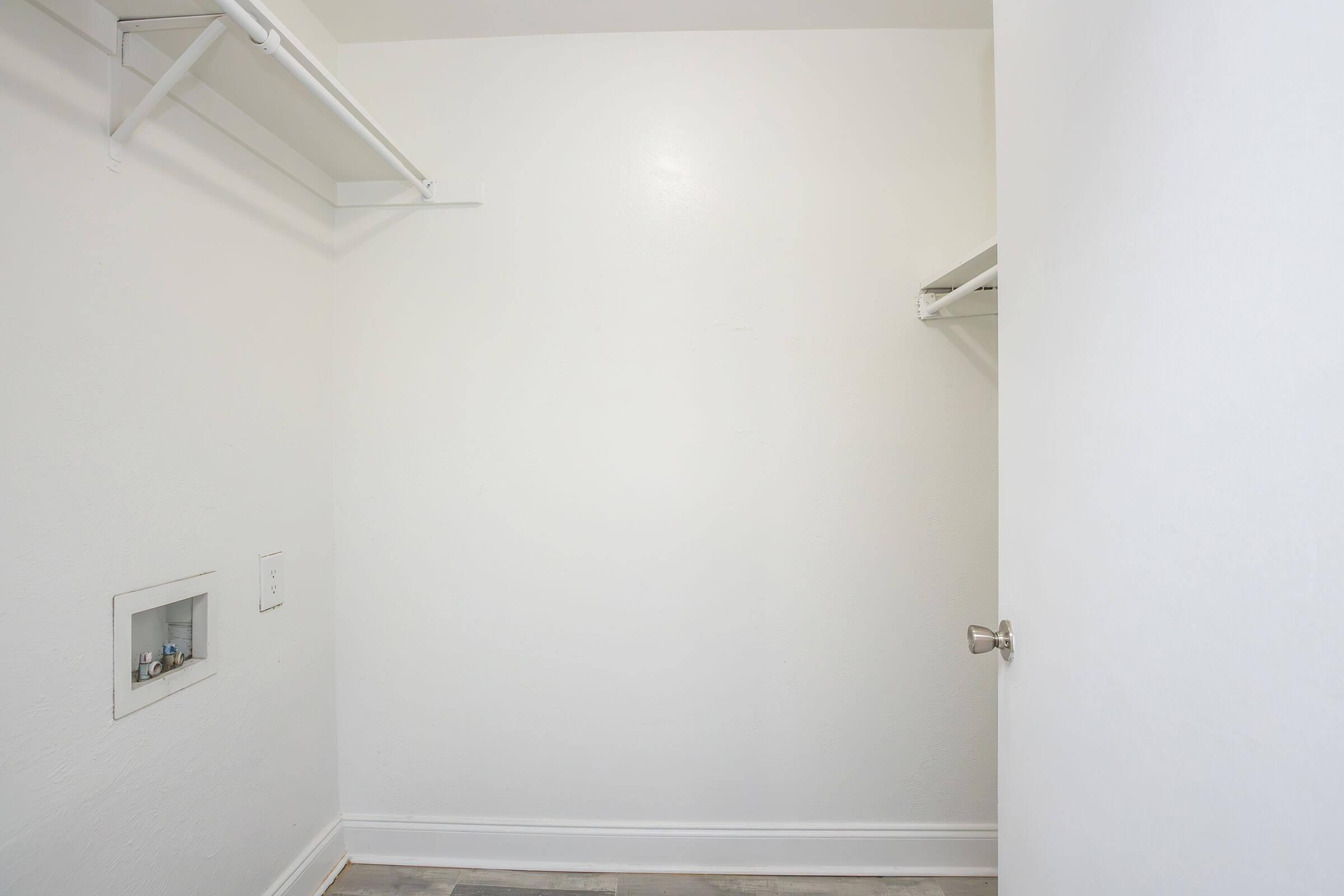 Empty closet with white walls and flooring. The closet features a single door on the right, a small opening on the back wall, and two shelves installed on the left side. The overall space appears bright and uncluttered.