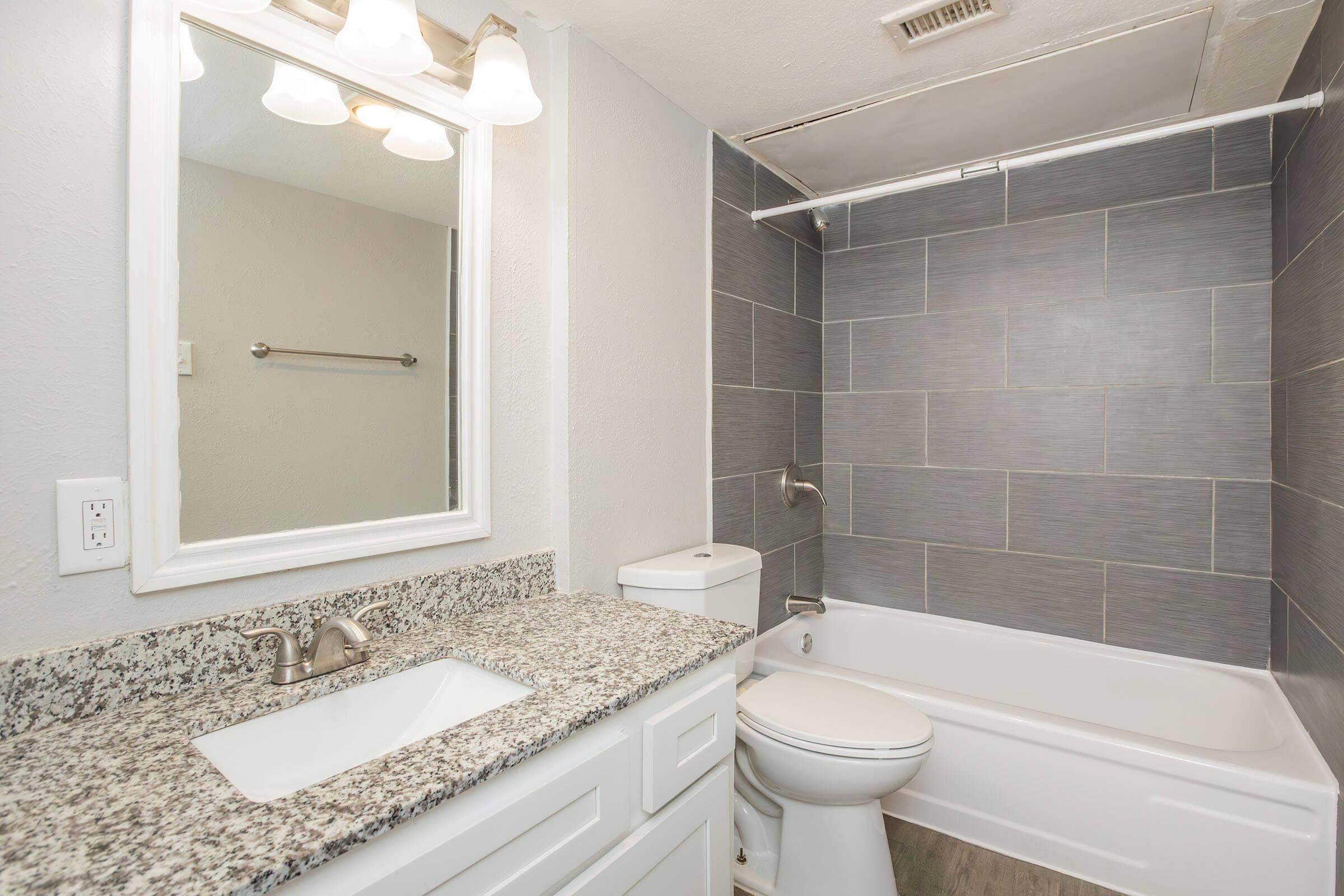 A modern bathroom featuring gray tiled walls, a white bathtub, a white toilet, and a granite countertop with an integrated sink. The lighting is bright with three fixtures above the mirror, providing a spacious and clean appearance.