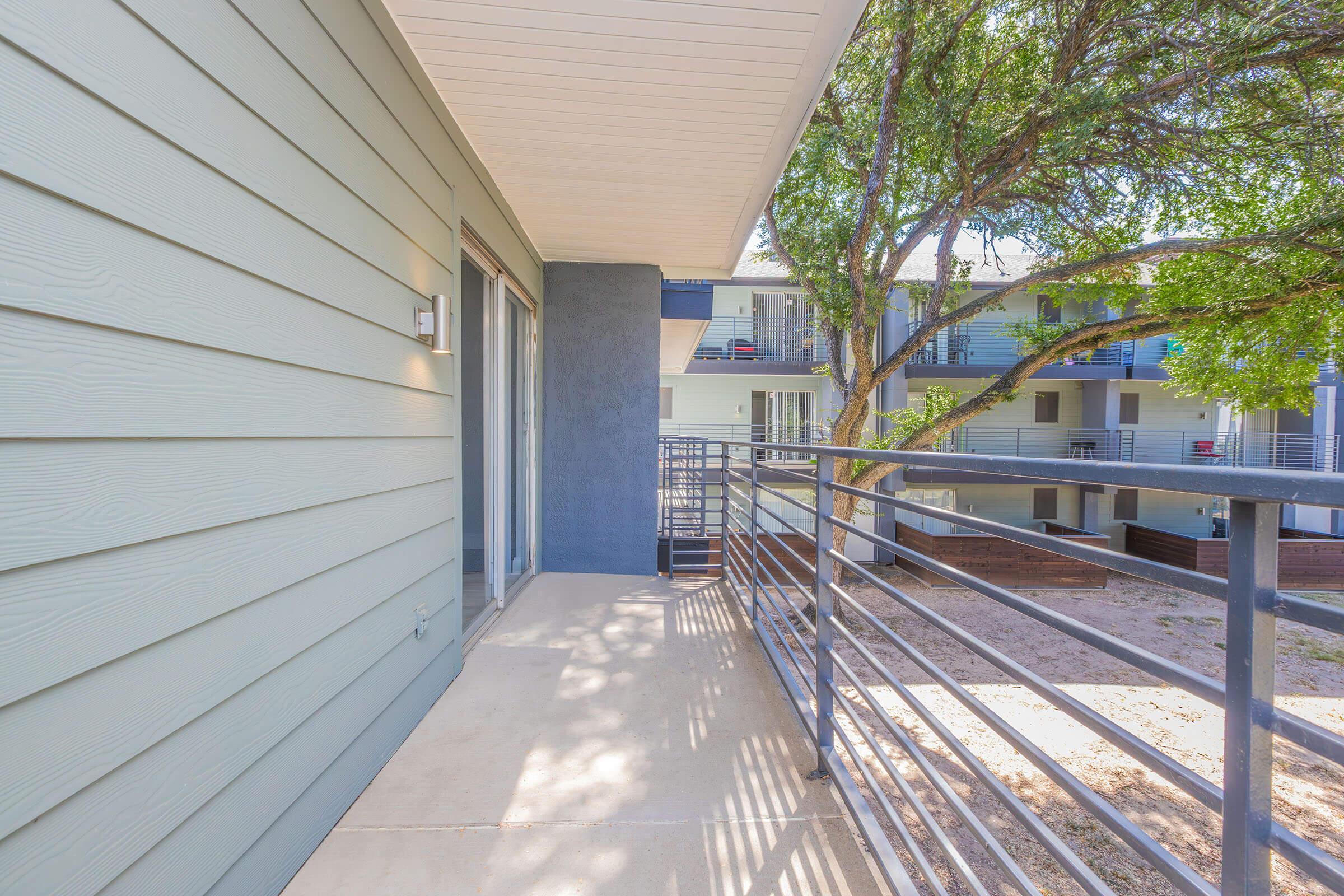 A balcony view with a minimalist design, featuring a concrete floor, metal railing, and green trees nearby. The background shows the exterior of an apartment building with multiple levels and balconies. Natural light brightens the space, creating a serene atmosphere.
