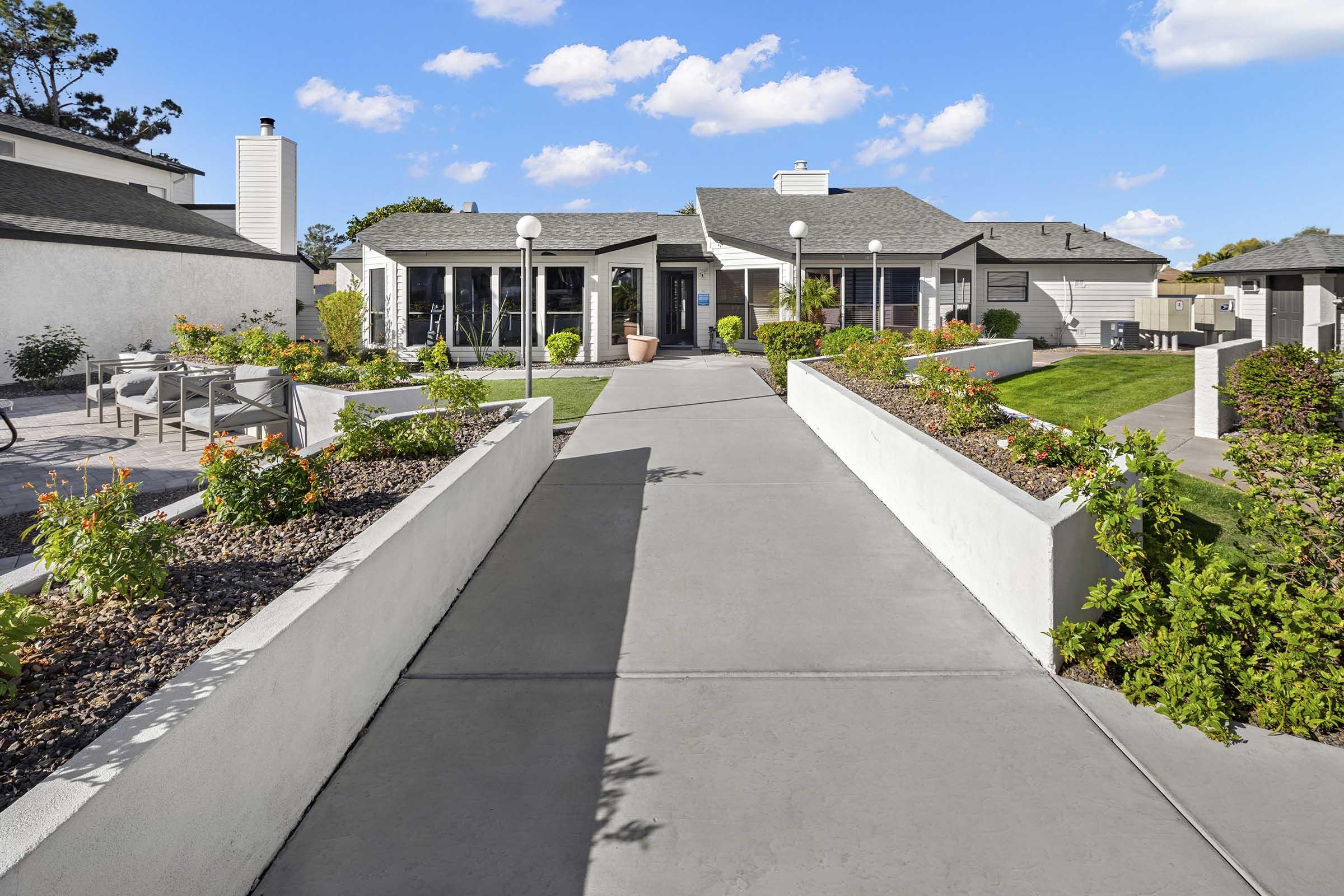 A neatly landscaped pathway leading to a modern single-story house with large windows. The pathway is flanked by blooming flower beds and green grass, while the sky is bright blue with fluffy white clouds. Outdoor seating areas are visible on the left, enhancing the inviting atmosphere of the property.
