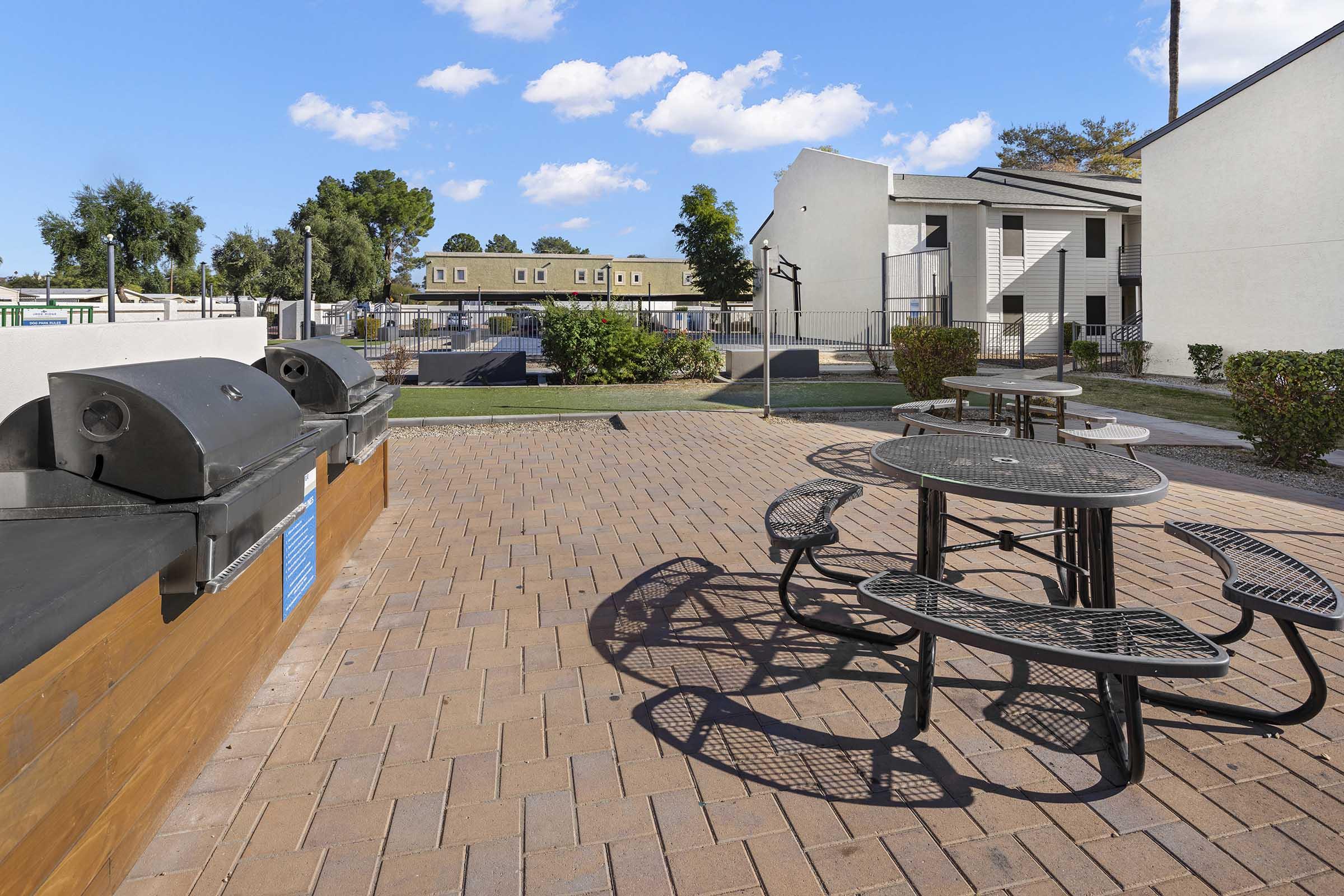 Outdoor area featuring several picnic tables and barbecue grills on a brick patio. In the background, there are green lawns and apartment buildings under a clear blue sky. The space is well-maintained, providing a community gathering spot.