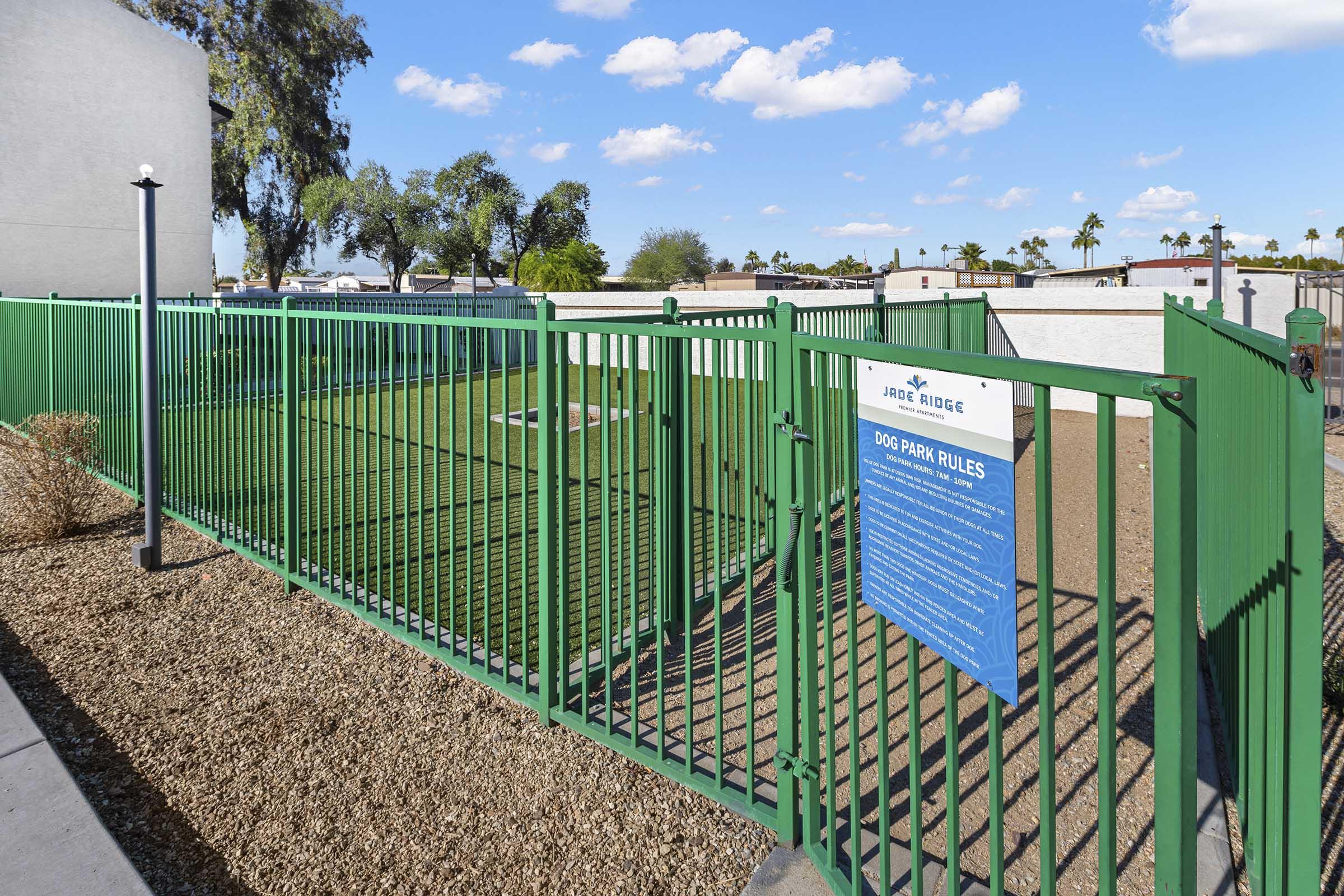A fenced dog park featuring green grass and gravel areas, with a sign listing park rules visible on the fence. The park is surrounded by trees and buildings, under a blue sky with scattered clouds.
