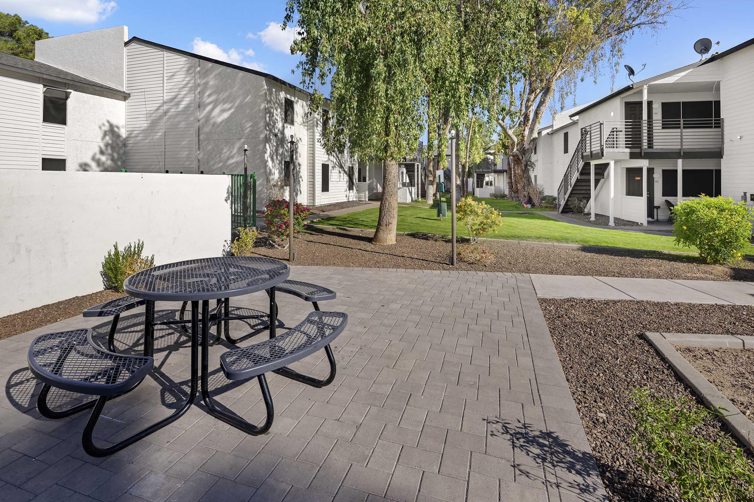 A landscaped courtyard featuring a circular picnic table surrounded by benches, with a well-maintained lawn, trees, and several apartment buildings in the background. The scene is bright and inviting, showcasing a pleasant outdoor living space.