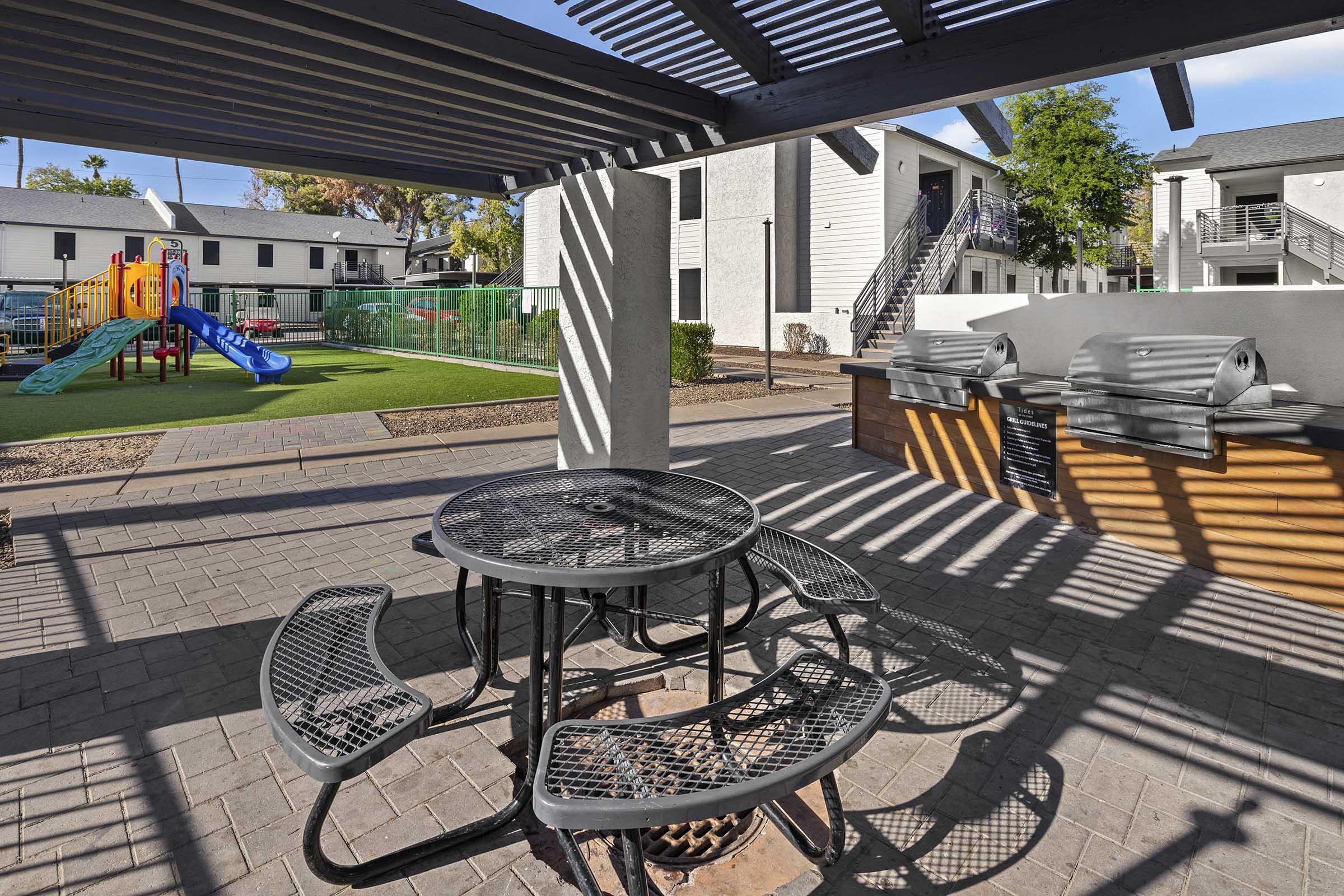 A shaded outdoor seating area with a black metal table and benches, overlooking a playground with colorful equipment and a grassy area. In the background, there are apartment buildings and a BBQ grill setup near the seating, under a pergola.