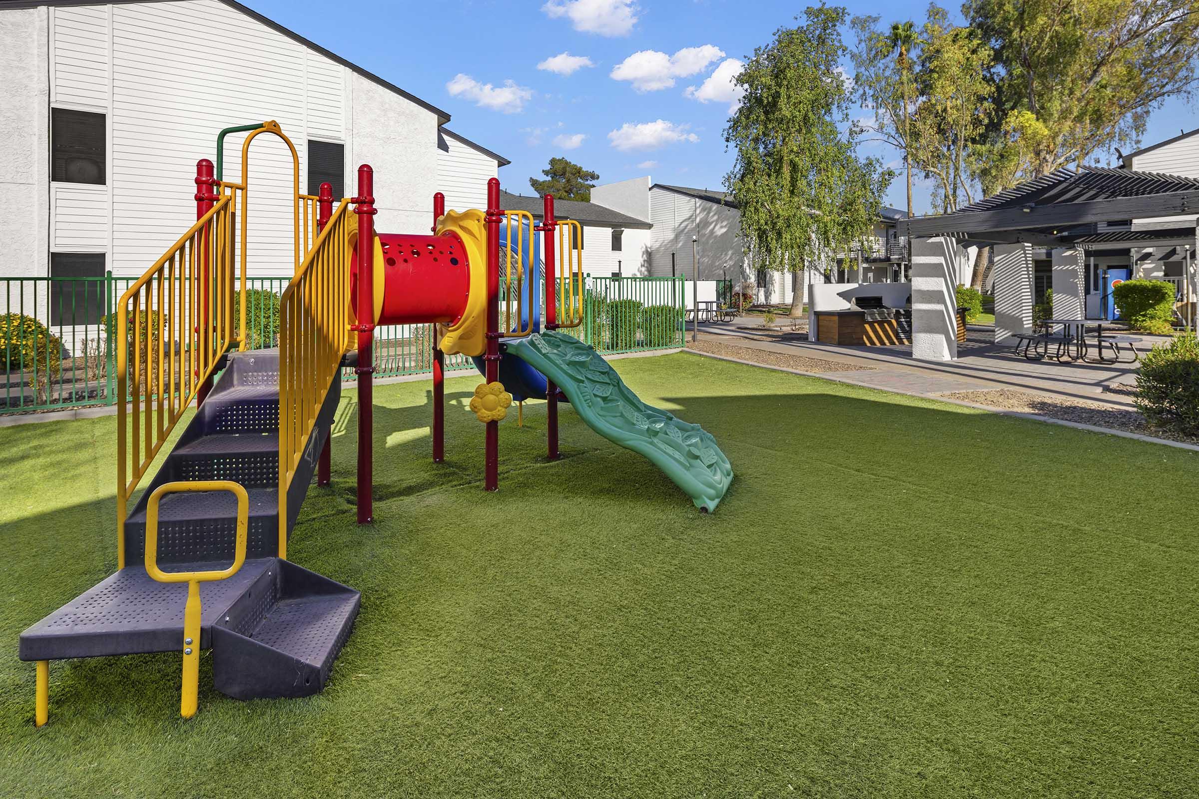 Colorful playground equipment with a slide and climbing structures, set on green artificial grass. Nearby, there's a shaded picnic area with tables, surrounded by trees and buildings in the background under a blue sky with fluffy clouds.