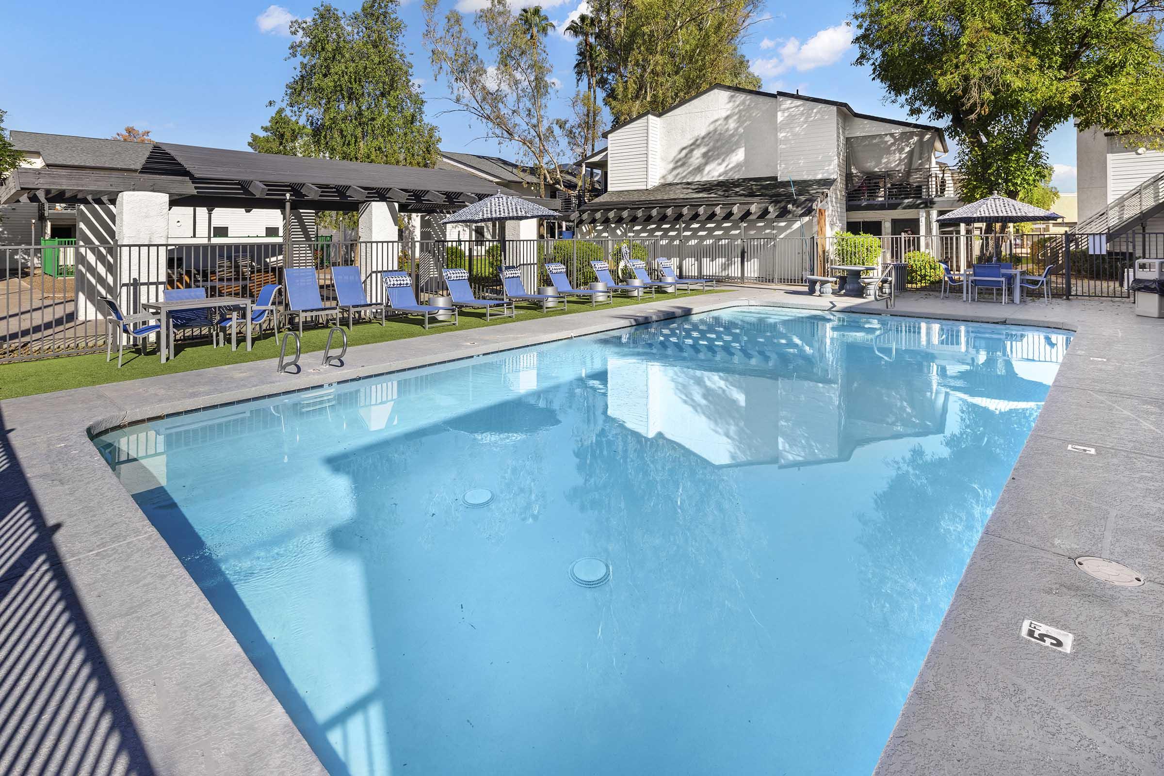 A clear blue swimming pool surrounded by lounge chairs and umbrellas. The pool area is well-maintained, featuring green grass and shaded seating. In the background, there are trees and residential buildings, creating a relaxing outdoor atmosphere.