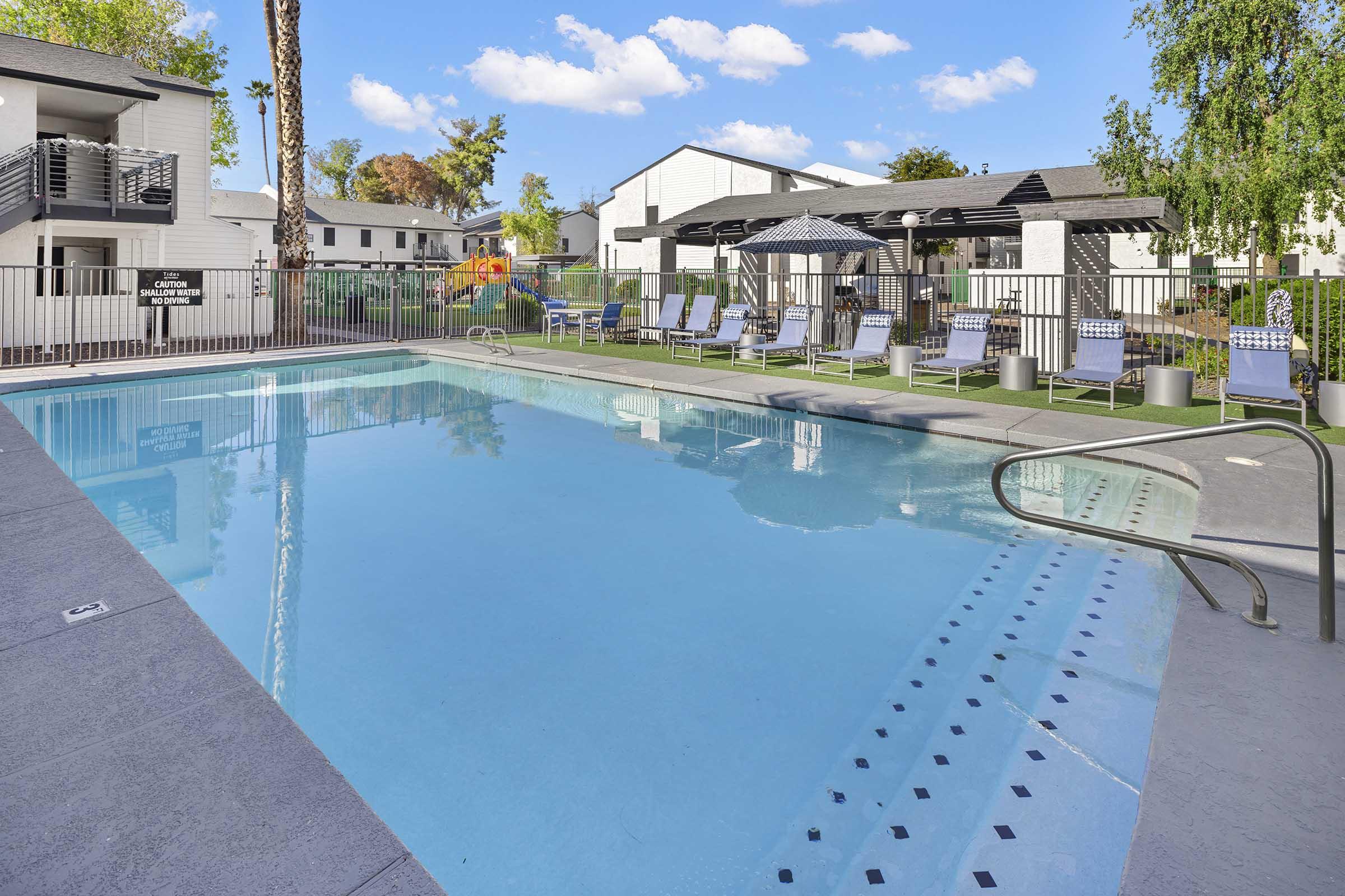 A clear swimming pool surrounded by lounge chairs and a fence. In the background, there are residential buildings with green lawns and trees. The sky is blue with a few clouds, adding to the inviting atmosphere of the outdoor space.