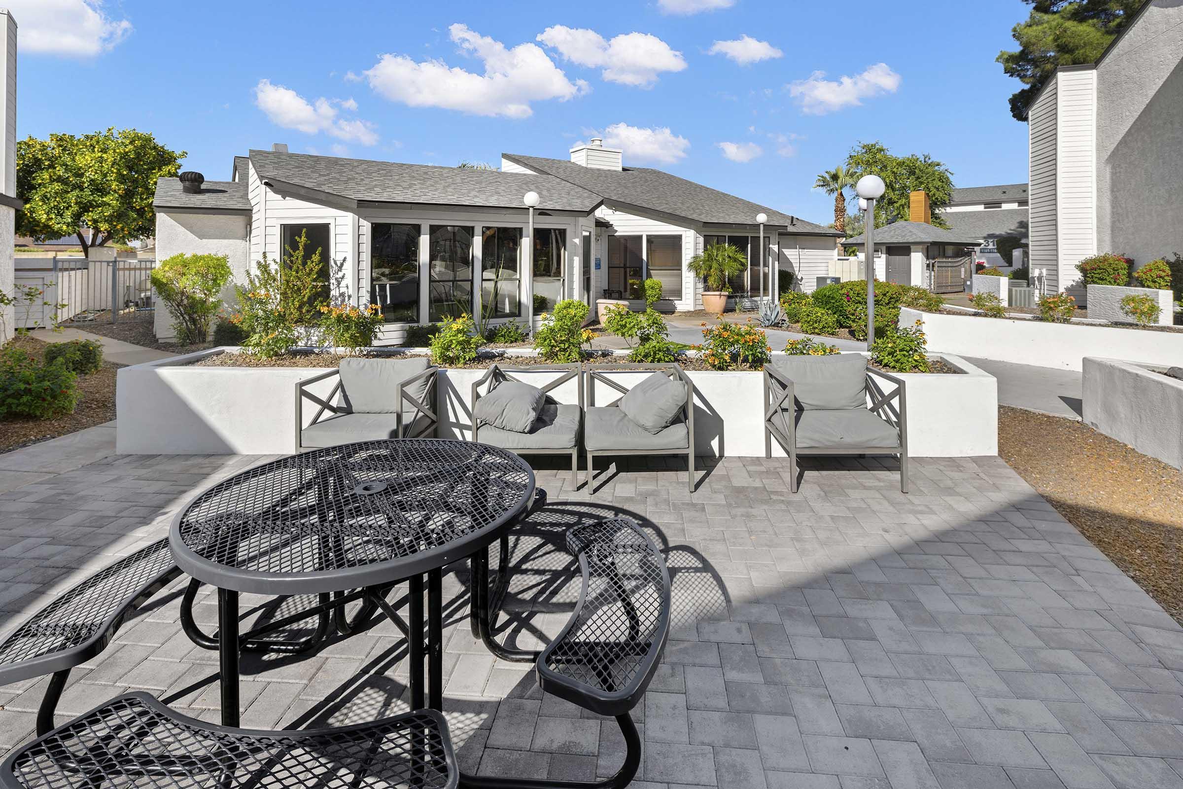 A spacious outdoor area featuring a round black metal dining table with several chairs, surrounded by landscaped gardens and pathways. In the background, there is a modern building with large windows, greenery, and decorative lighting, under a clear blue sky with a few fluffy clouds.
