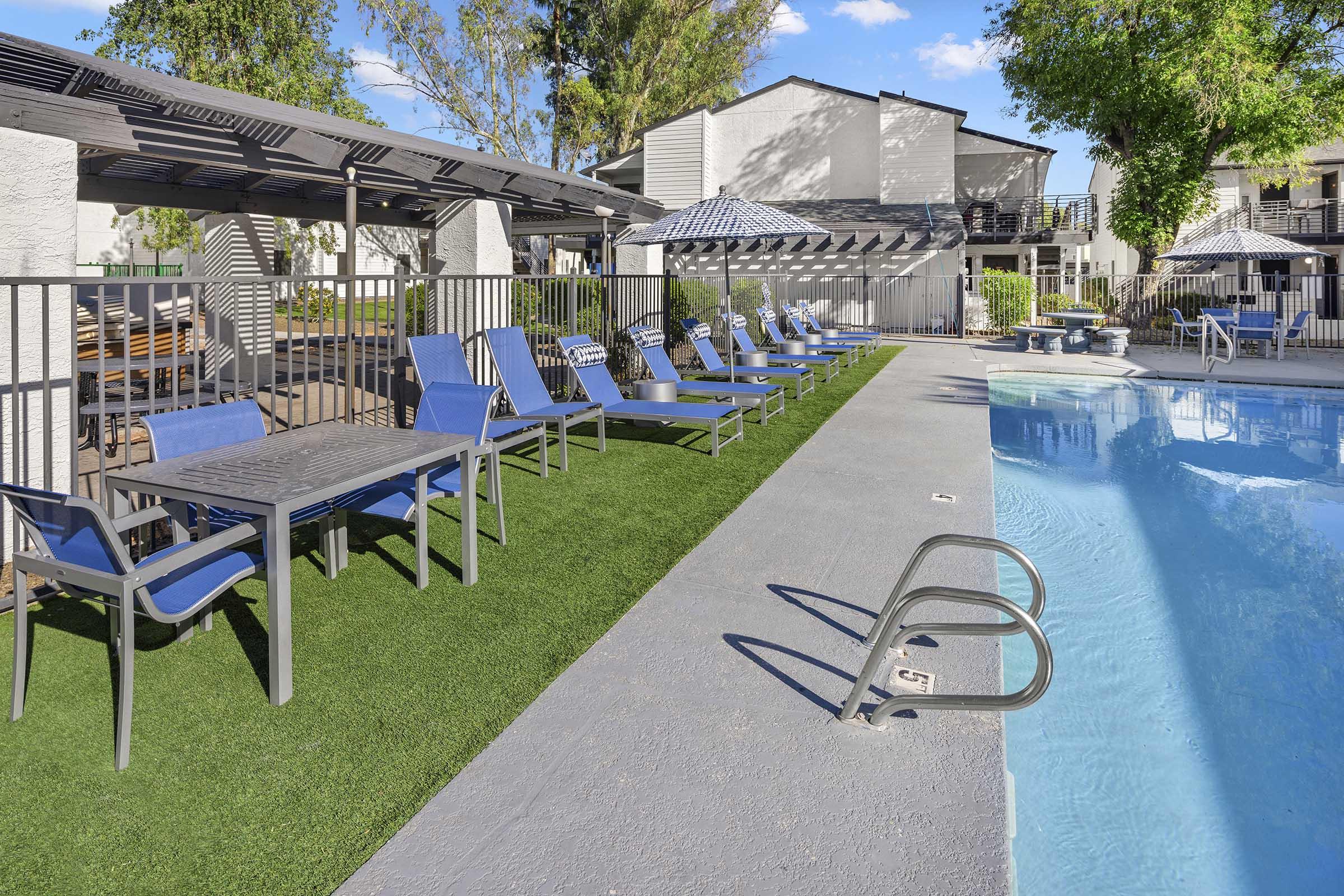 A spacious poolside area featuring a sparkling blue pool, lounge chairs, and shaded tables. The surroundings are landscaped with green grass and the sun is shining, creating a relaxing atmosphere. Several umbrellas provide shade, enhancing the inviting outdoor space for leisure and relaxation.