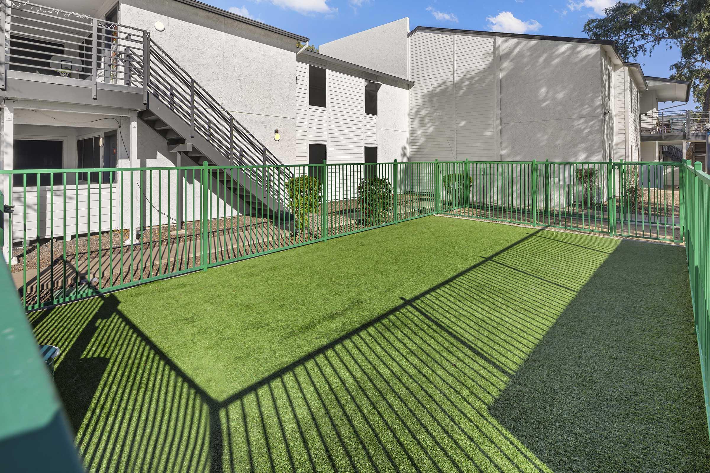 A fenced grassy area with artificial turf, surrounded by green fencing. The background shows a multi-story building with stairs and an exterior walkway. Sunlight creates shadows on the turf, highlighting the green space in an apartment complex setting.