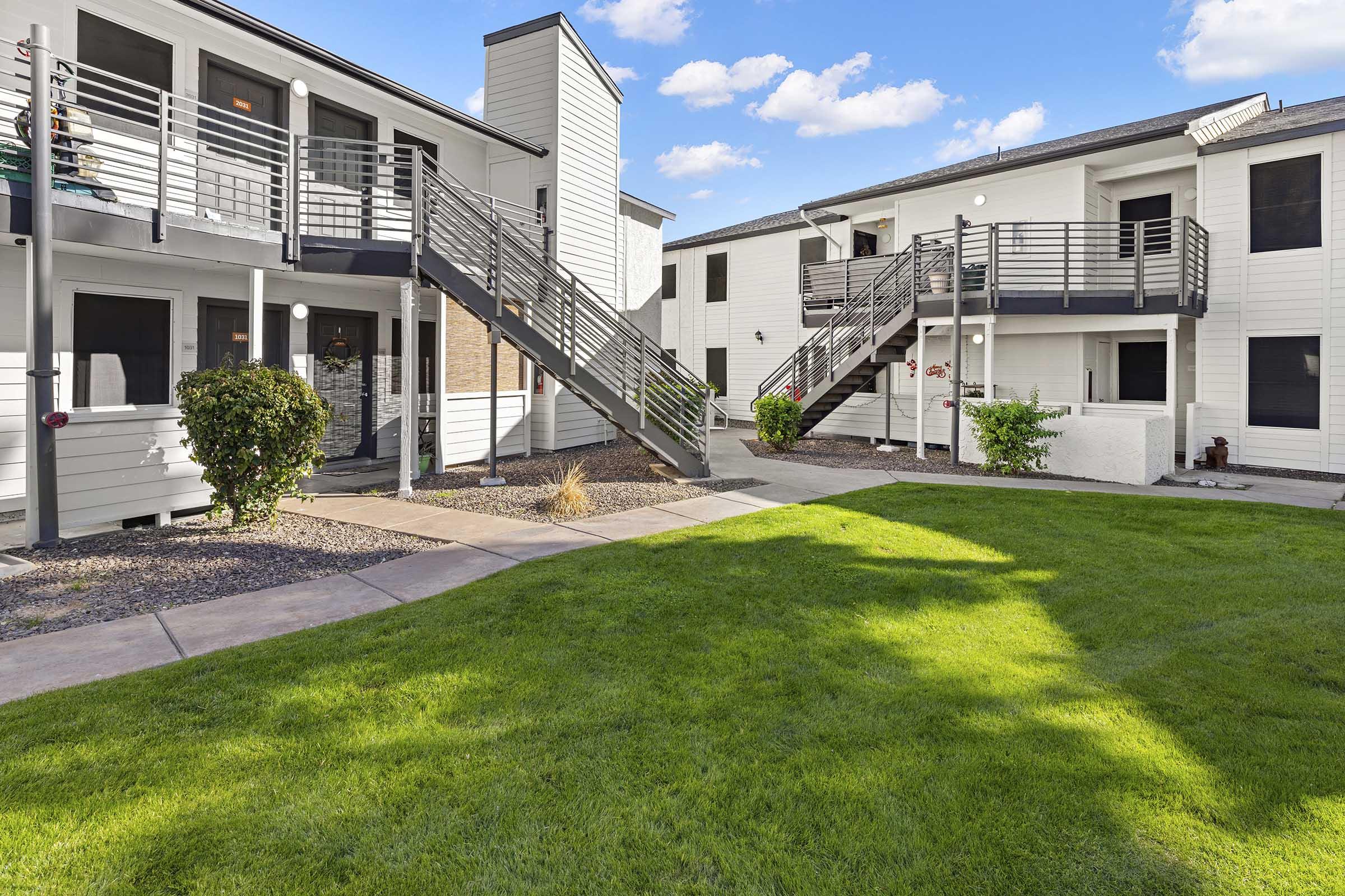 View of a landscaped courtyard surrounded by two-story apartment buildings. The scene features green grass, paved walkways, and stairs leading to upper-level entrances. The sky is partly cloudy, providing a bright and inviting atmosphere.