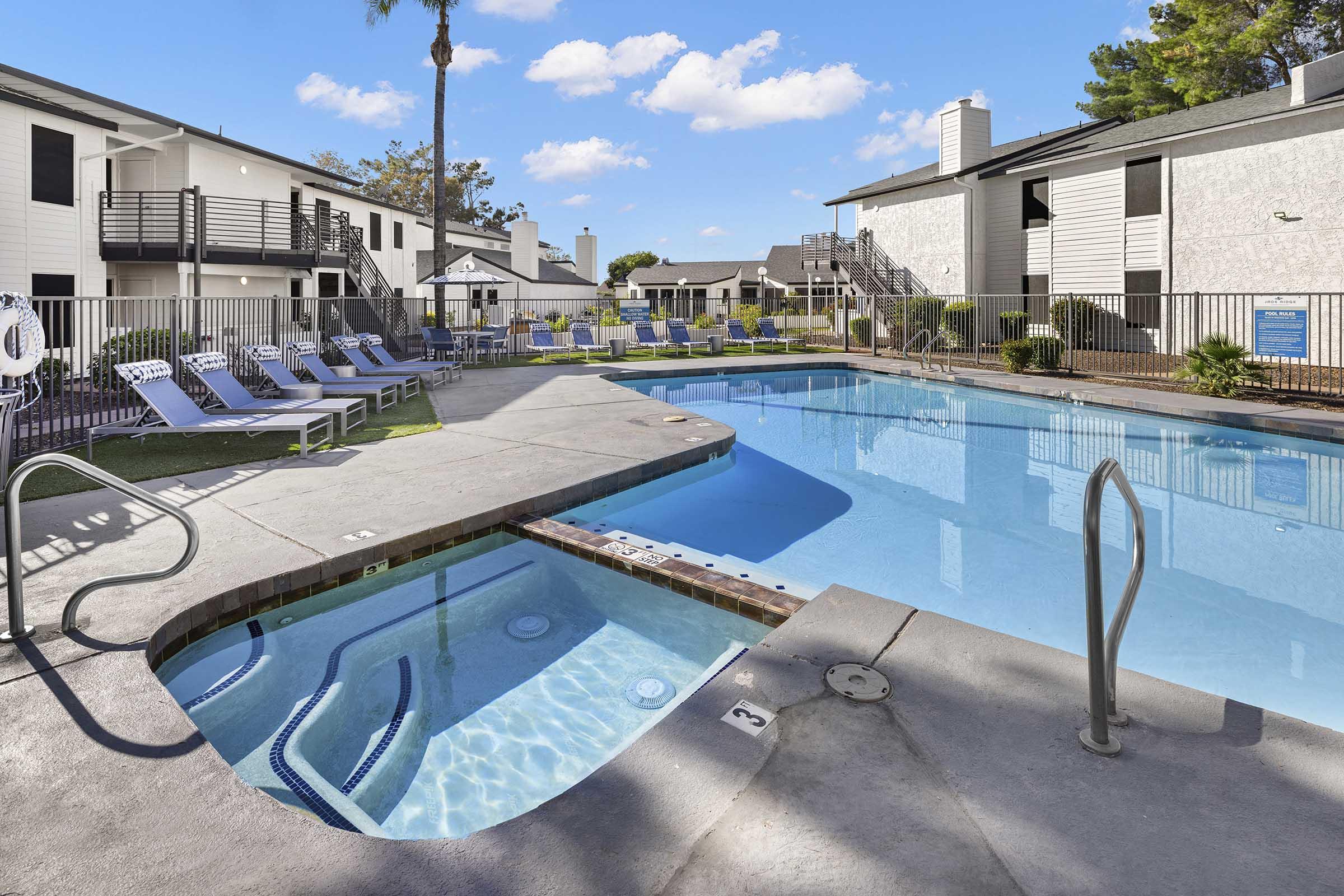 A clear blue swimming pool surrounded by lounge chairs and a hot tub. The pool area is framed by white apartment buildings, with palm trees and a bright blue sky in the background. The scene conveys a relaxing outdoor atmosphere.