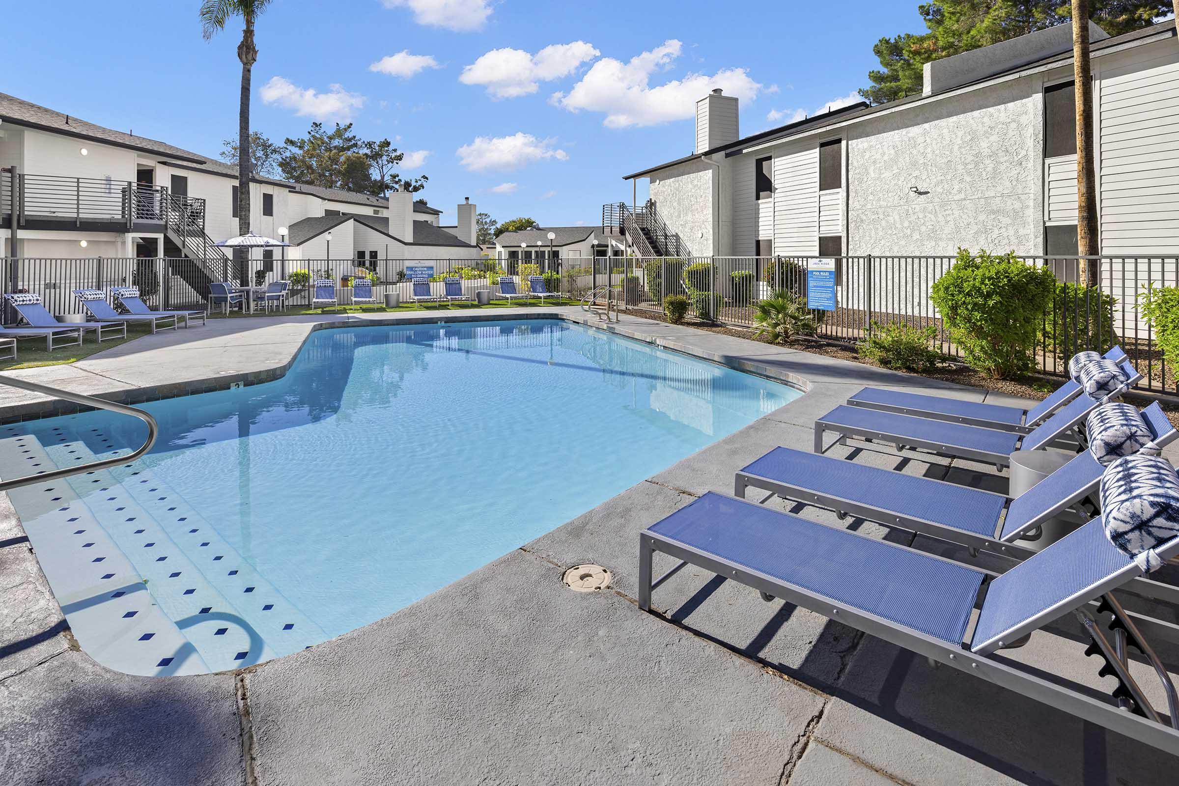 A clear blue swimming pool surrounded by lounge chairs in a residential complex. The pool area features green landscaping, a fence, and nearby buildings under a bright blue sky with fluffy clouds. Perfect for relaxation and leisure activities.