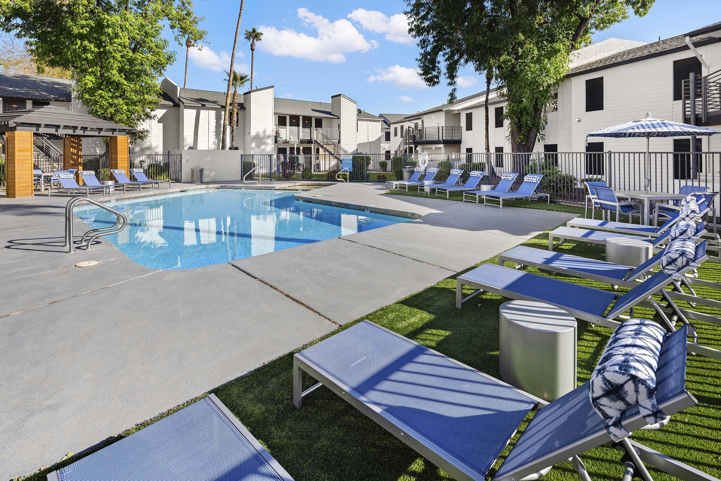 A sparkling blue swimming pool surrounded by lounge chairs on a sunny day. A few umbrellas provide shade, with green grass and trees in the background. Several apartment buildings are visible, creating a relaxed and inviting atmosphere.