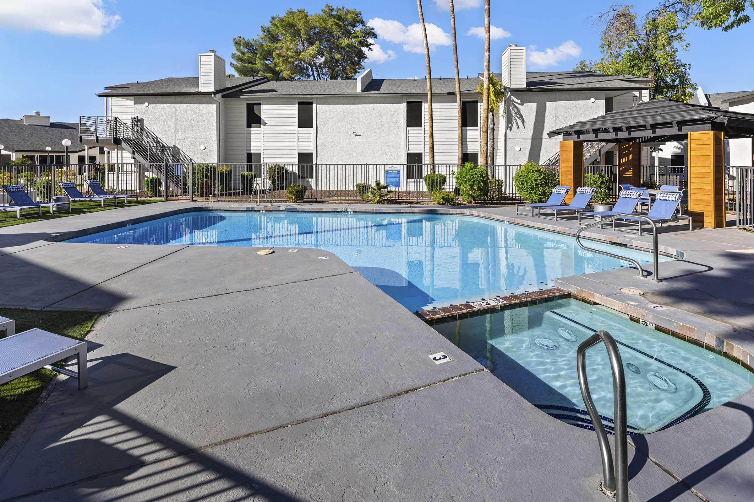 A large swimming pool surrounded by lounge chairs and greenery, with a hot tub nearby. The pool area features a clean concrete deck, palm trees, and a modern shaded gazebo. Bright blue sky with a few clouds in the background. Ideal for relaxation and leisure activities.