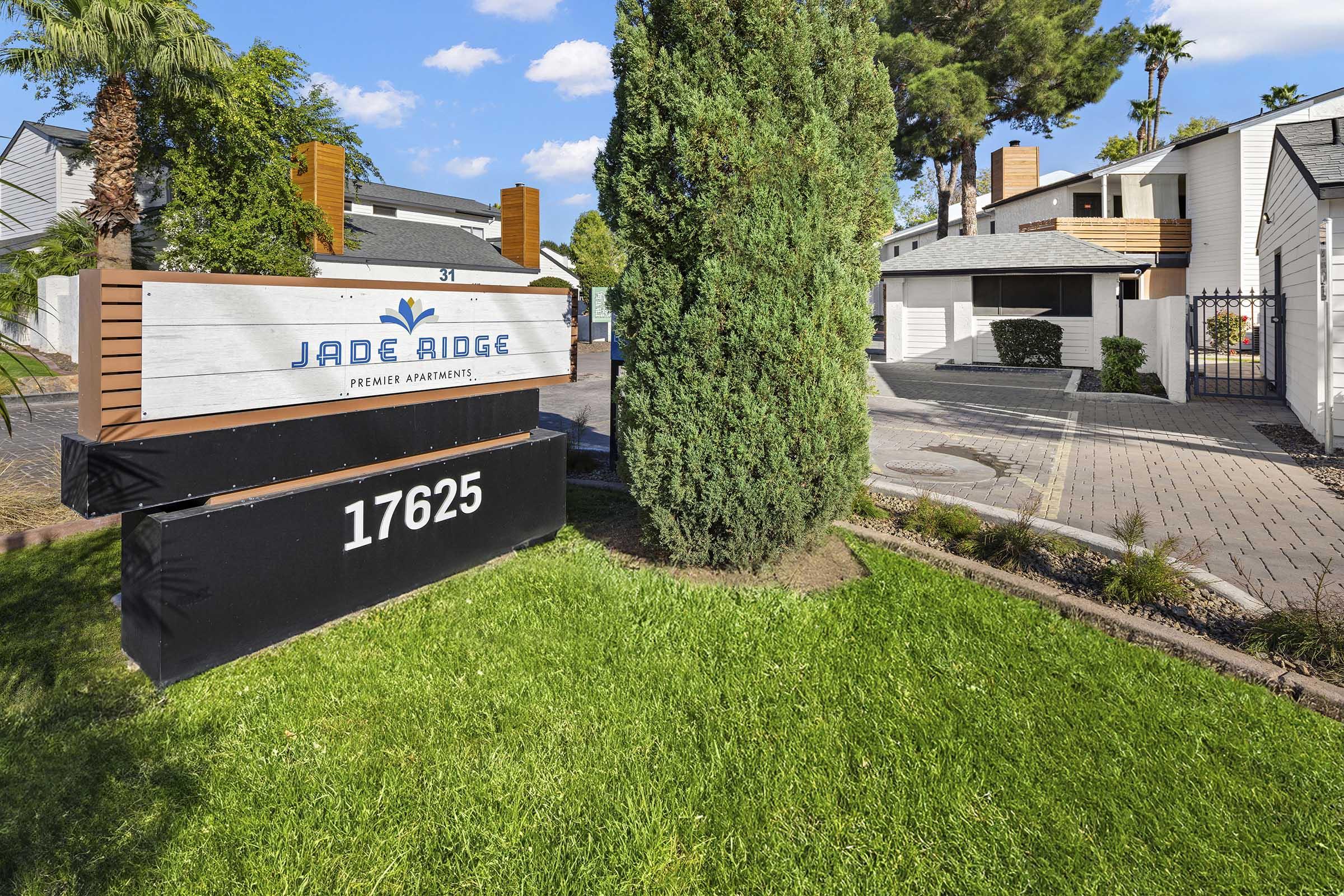 Sign for Jade Ridge Premier Apartments, featuring the name prominently displayed. The sign is situated in a well-maintained grassy area with shrubs and palm trees in the background. A pathway leads to the apartments visible in the distance, under a bright blue sky with clouds.