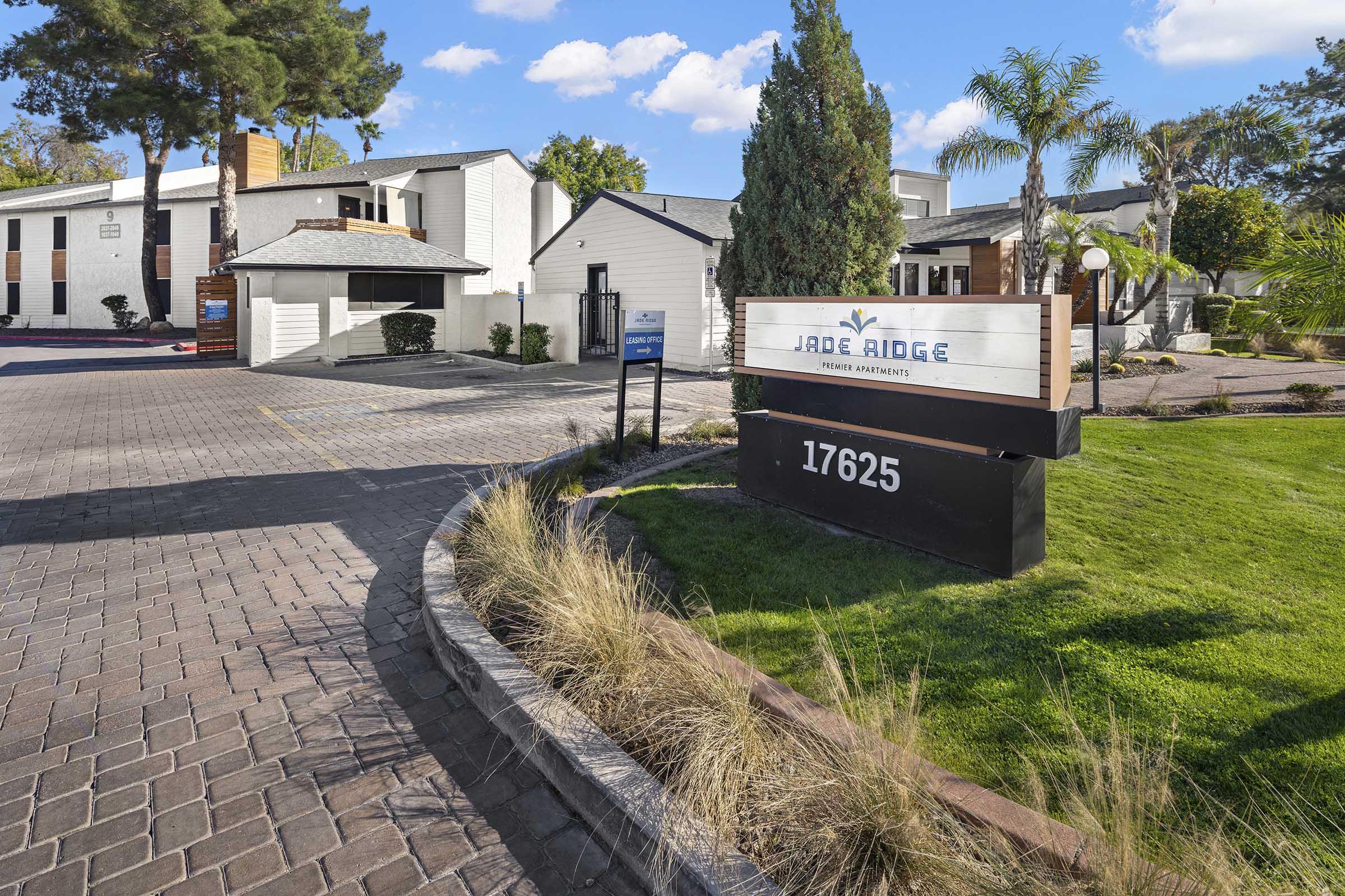 Sign for Jade Ridge Private Apartments at the entrance, with the address number 17625. The scene features landscaped grounds with palm trees and a clear blue sky. Buildings are visible in the background, along with a paved pathway leading to the entrance.