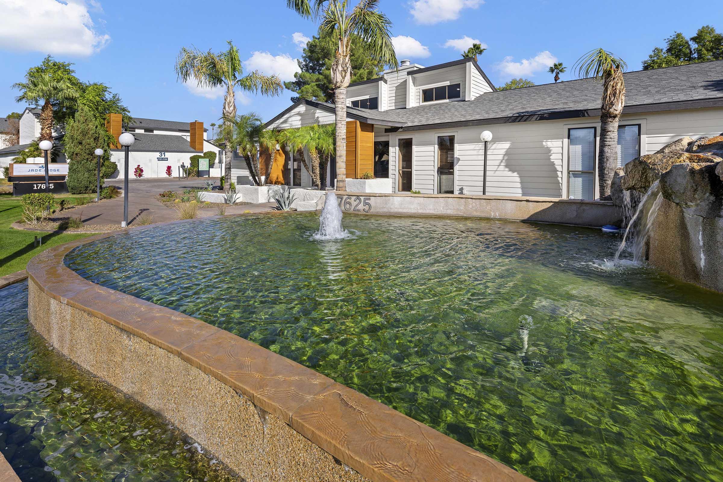 A tranquil water feature with flowing fountains, reflecting palm trees and a modern building. The scene includes a clear green pond surrounded by landscaped areas, creating a peaceful outdoor environment. Bright blue sky with a few clouds completes the serene atmosphere.