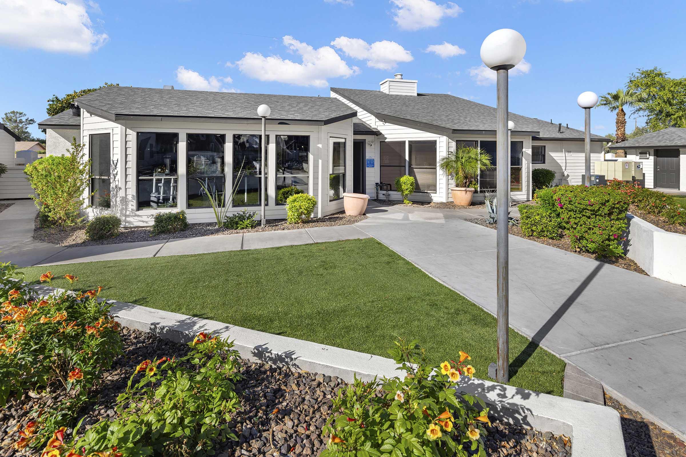 A well-maintained residential building with large windows, surrounded by landscaped gardens featuring colorful flowers. The pathway is lined with modern light fixtures, leading to a green lawn. The sky is clear with a few clouds, creating a bright and inviting atmosphere.