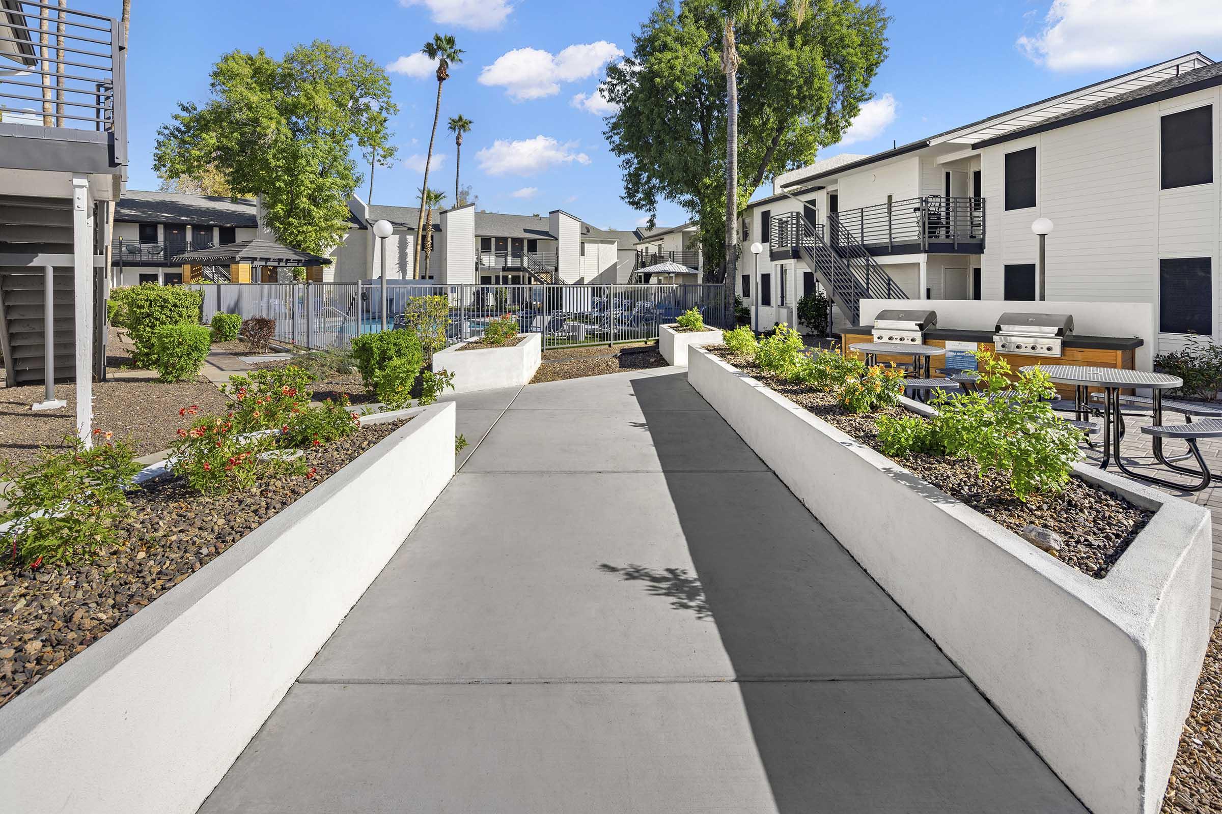 Pathway surrounded by landscaped gardens, leading through a residential complex with seating areas and barbecues. Various trees and plants enhance the outdoor space under a bright blue sky with a few clouds. Apartment buildings are visible in the background, indicating a well-maintained living environment.