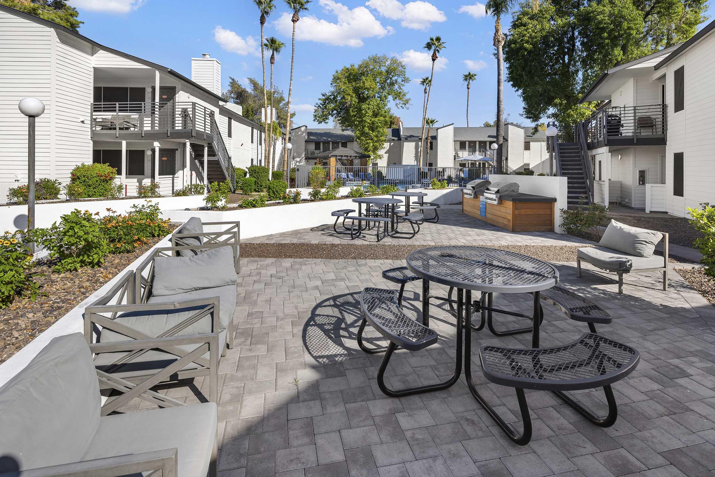 Outdoor seating area in a modern apartment complex featuring tables and chairs surrounded by landscaped greenery, palm trees, and multiple buildings in the background. The space is designed for relaxation and social gatherings, with a bright blue sky overhead.