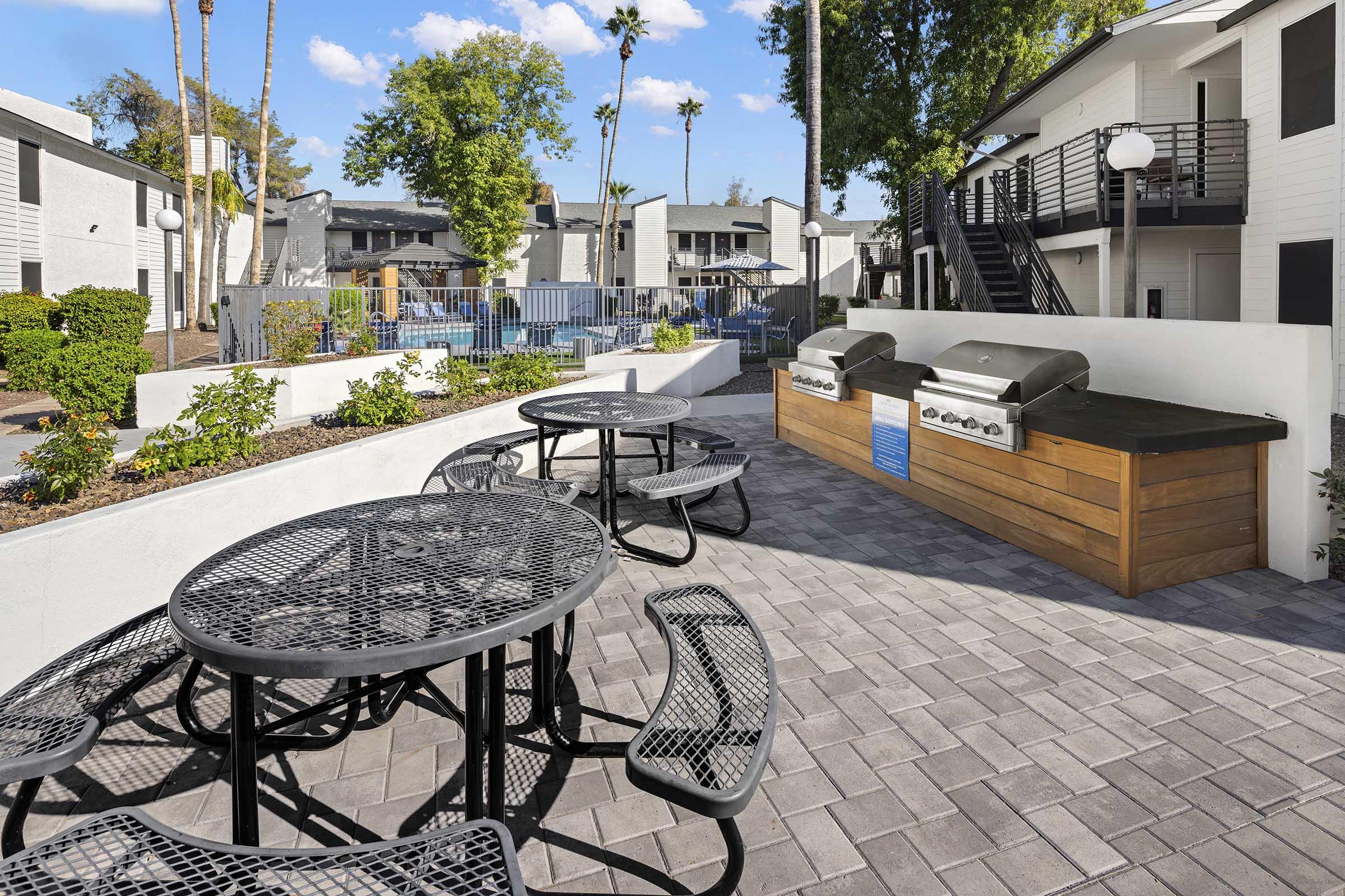 Outdoor grill area in an apartment complex, featuring multiple picnic tables and barbecue grills. The space is surrounded by green landscaping, palm trees, and a swimming pool in the background, creating a welcoming environment for residents to gather and enjoy outdoor cooking and dining.