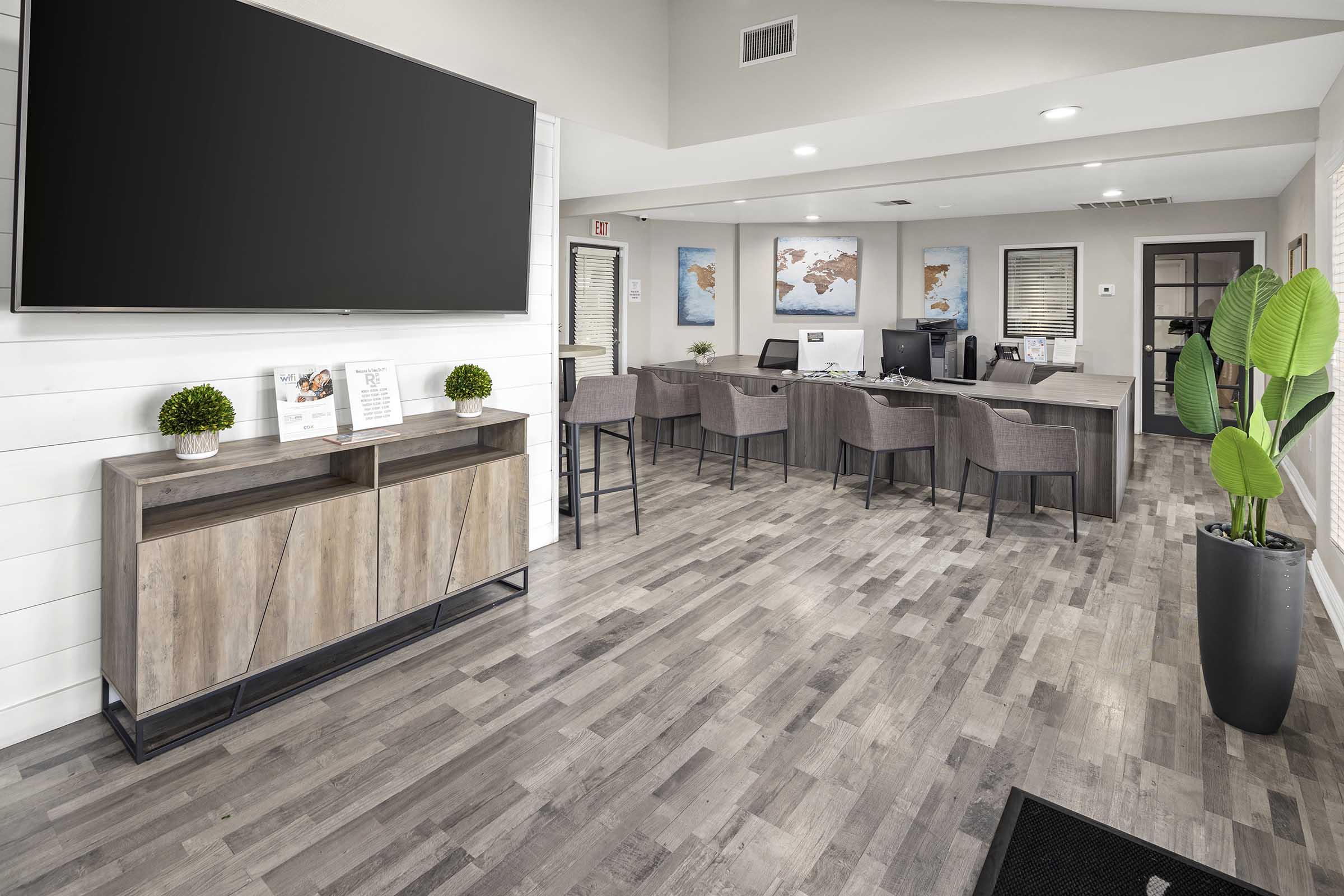 Interior view of a modern office reception area featuring a large flat-screen TV on the wall, a wooden console table with decorative plants, several gray chairs, and multiple desks with computers. Light-colored walls and artwork depicting maps enhance the spacious and welcoming atmosphere.
