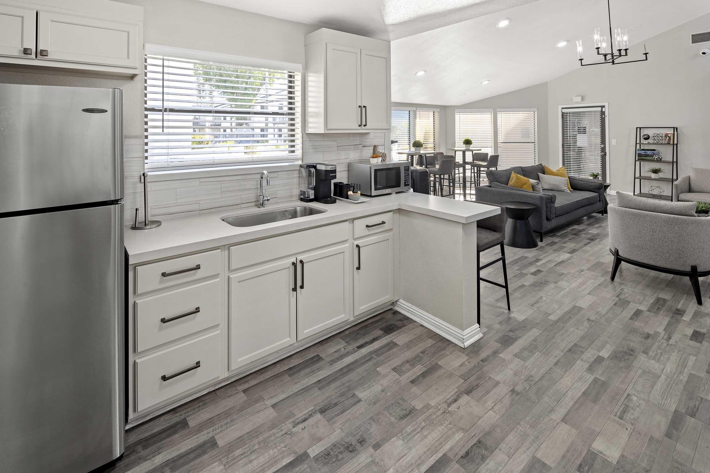 A modern kitchen with stainless steel appliances, including a refrigerator and microwave, paired with white cabinets and a sink. The open space features grey wooden flooring, a dining area with a table and chairs, and a cozy living room with grey sofas and colorful cushions, illuminated by natural light from large windows.