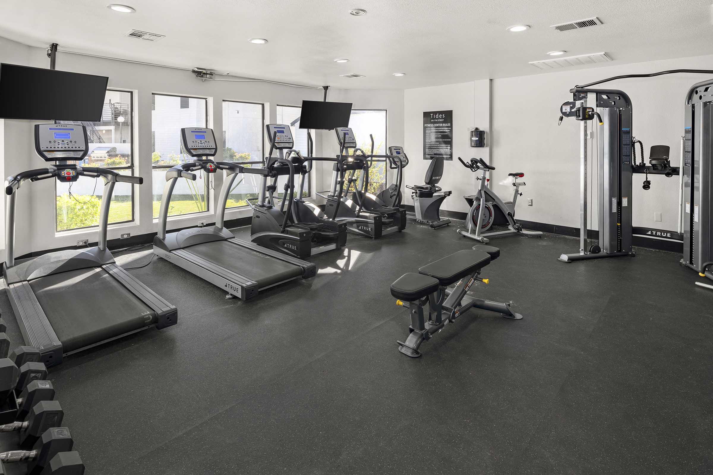 A modern gym featuring treadmills, stationary bikes, an adjustable bench, and weight dumbbells. The space is well-lit with large windows and two mounted televisions. Fitness equipment is arranged neatly on a black rubber floor, creating a clean and inviting workout environment.