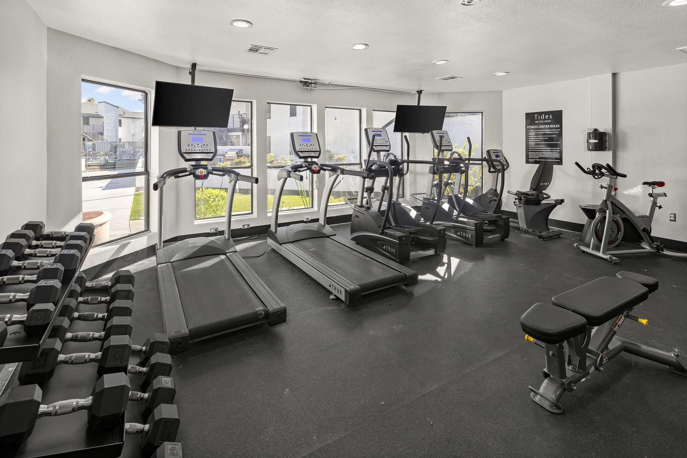 A modern gym interior featuring multiple treadmills, exercise bikes, and a set of dumbbells. Large windows allow natural light to enter, and two flat-screen TVs are mounted on the walls. The flooring is dark, enhancing the contemporary design of the workout space.
