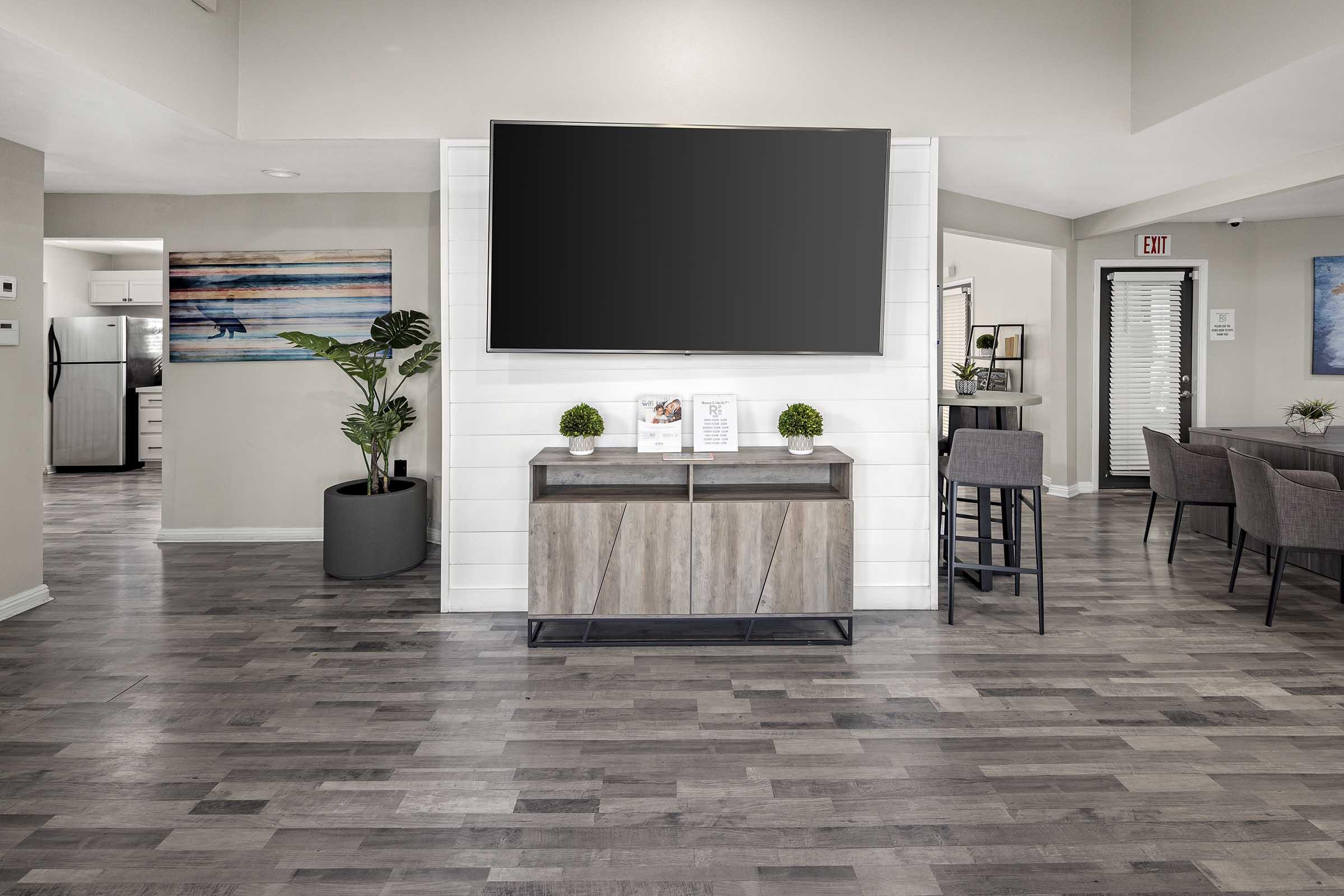 Modern interior of a spacious living area featuring a large wall-mounted television, a stylish wooden cabinet with decorative items, a potted plant, and a dining area with gray chairs. Soft, neutral colors and wooden flooring create a welcoming atmosphere.