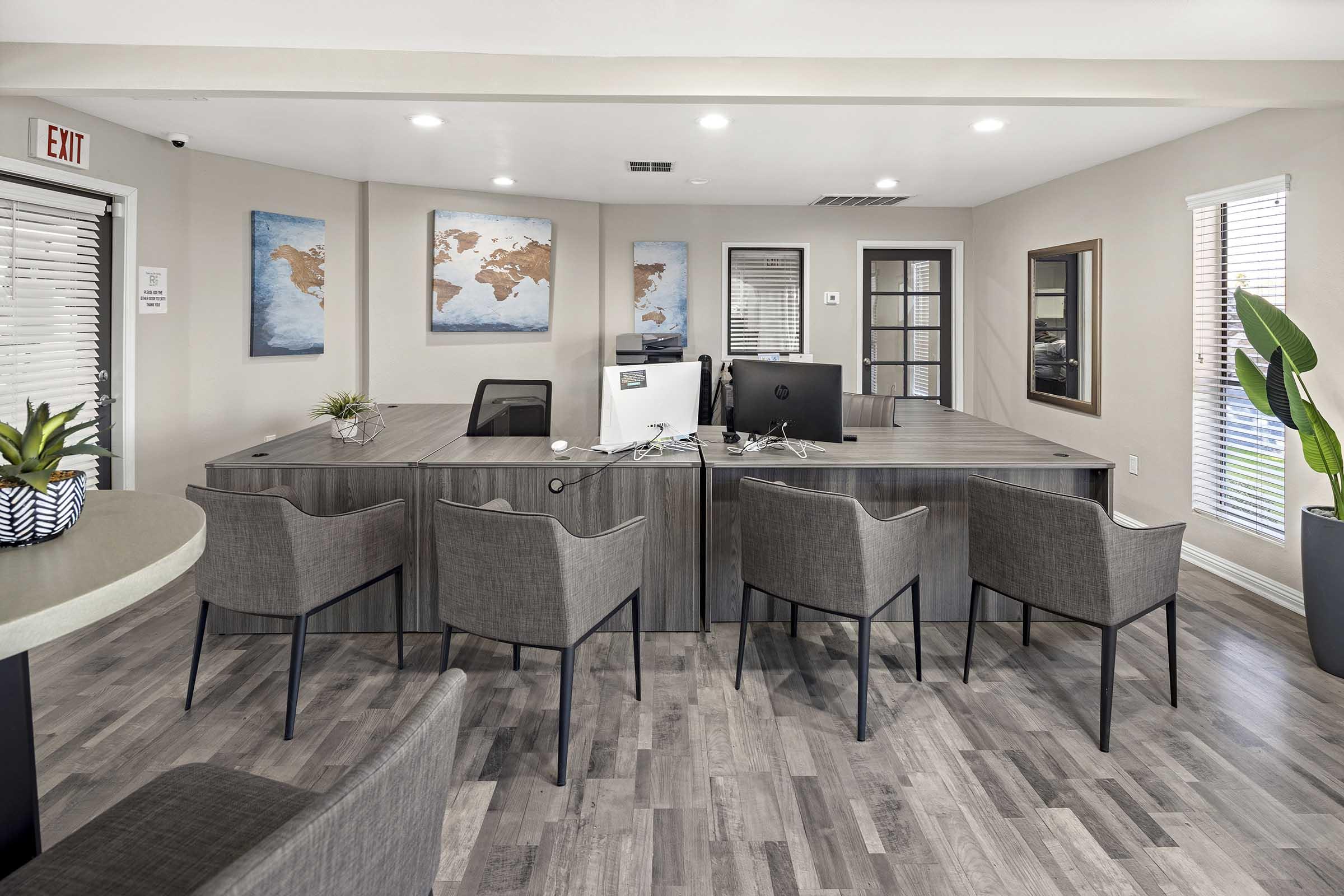 A modern office or reception area featuring a large wooden desk with two computers, several gray chairs, and framed maps on the walls. The space is well-lit with natural light from windows, and decorative plants add a touch of greenery.