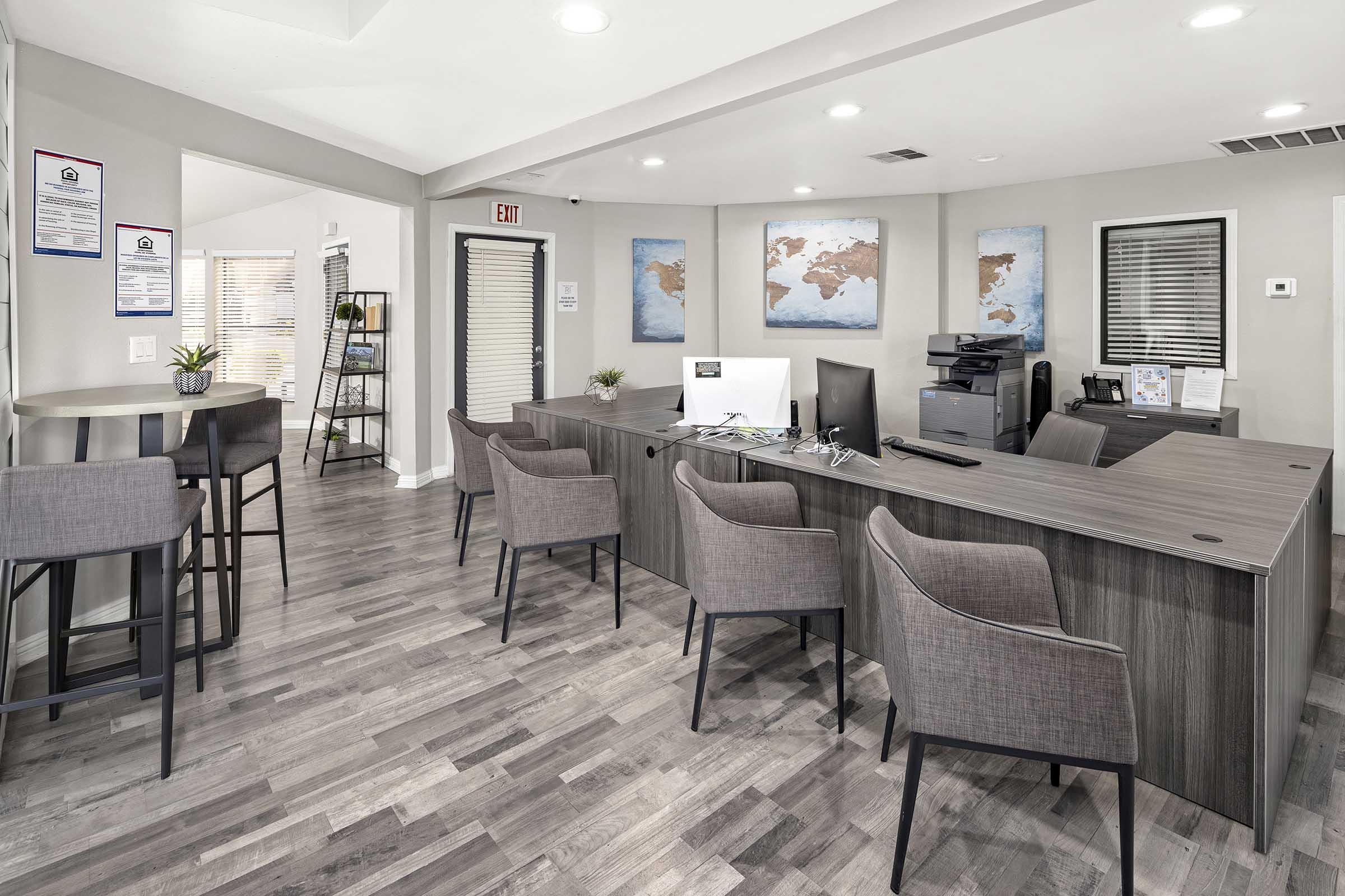Modern office interior featuring a reception area with a large wooden desk, computer, and printer. There are several gray chairs arranged around a small table, and decorative plants on the desk. The walls display maps of the world, and large windows provide natural light.