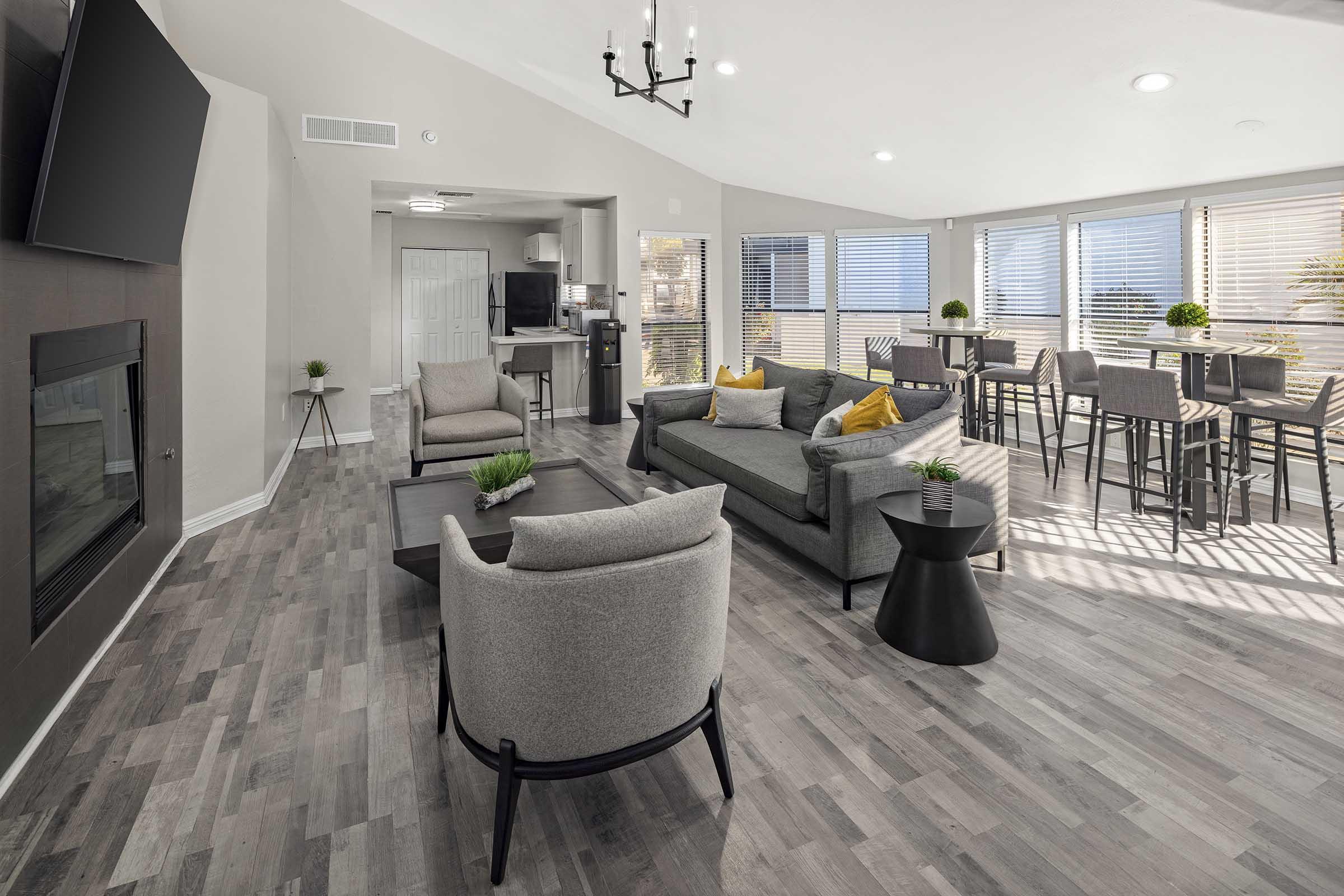 A modern living space featuring a comfortable seating area with gray sofas and accent pillows, a coffee table, and a fireplace. Large windows allow natural light, illuminating the room with a dining table and chairs in the background, creating a welcoming atmosphere. Hardwood flooring enhances the contemporary design.